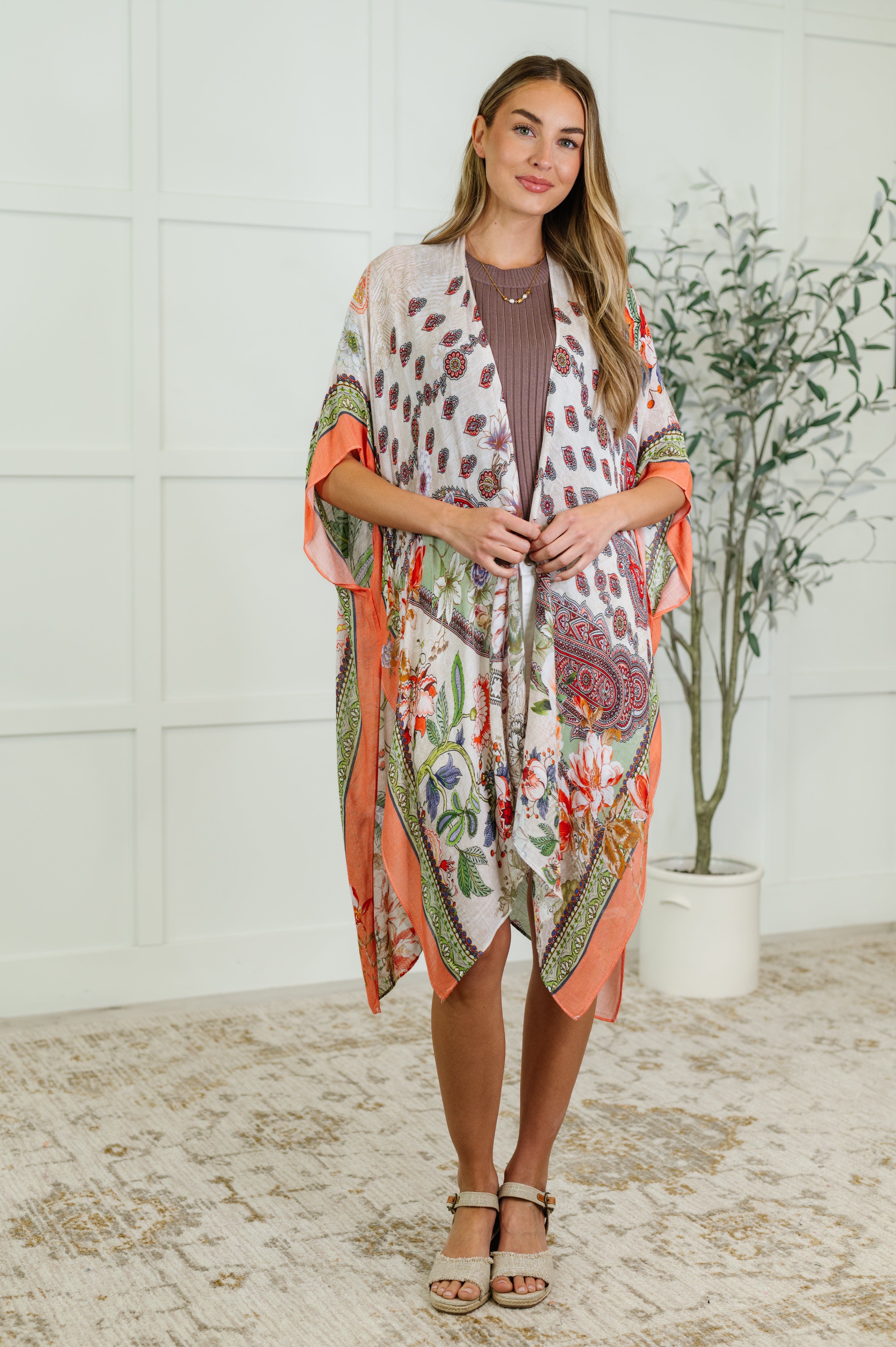 Smiling woman wearing an orange and green patterned kimono over a brown dress standing indoors near a plant