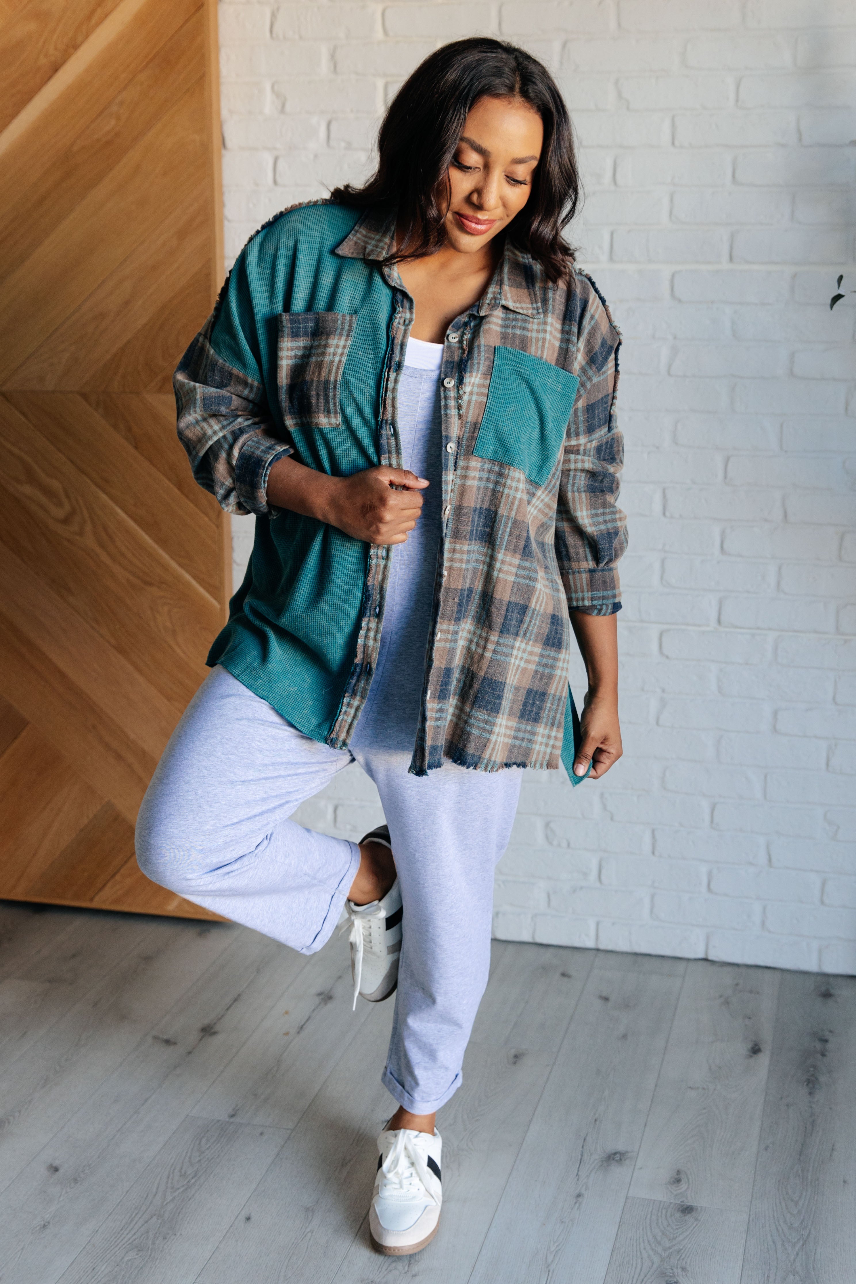 Woman wearing a teal and plaid oversized thermal shirt with pockets paired with light gray sweatpants and white sneakers indoors