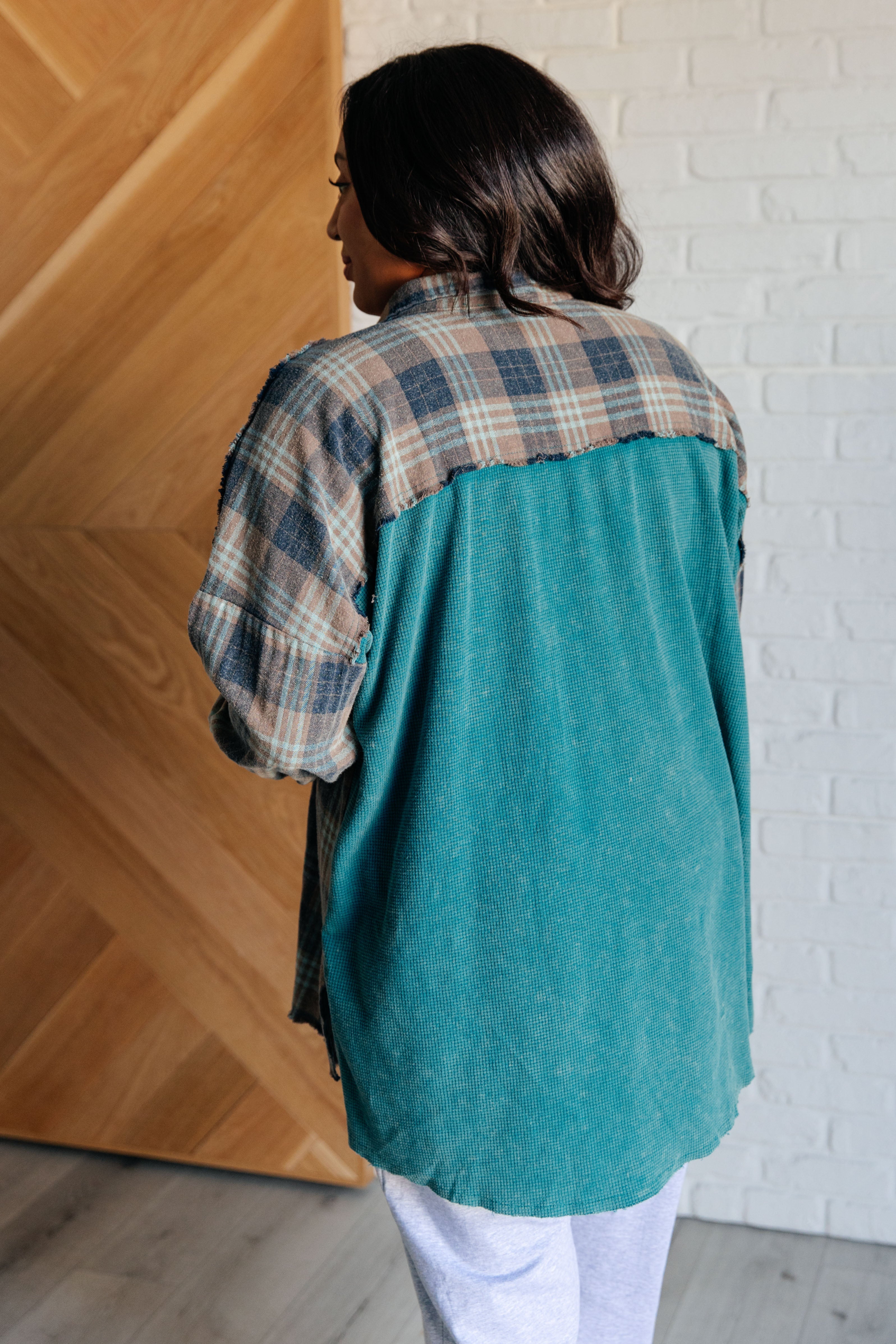 Woman wearing a teal and plaid thermal button-up shirt with raw edges standing indoors facing away
