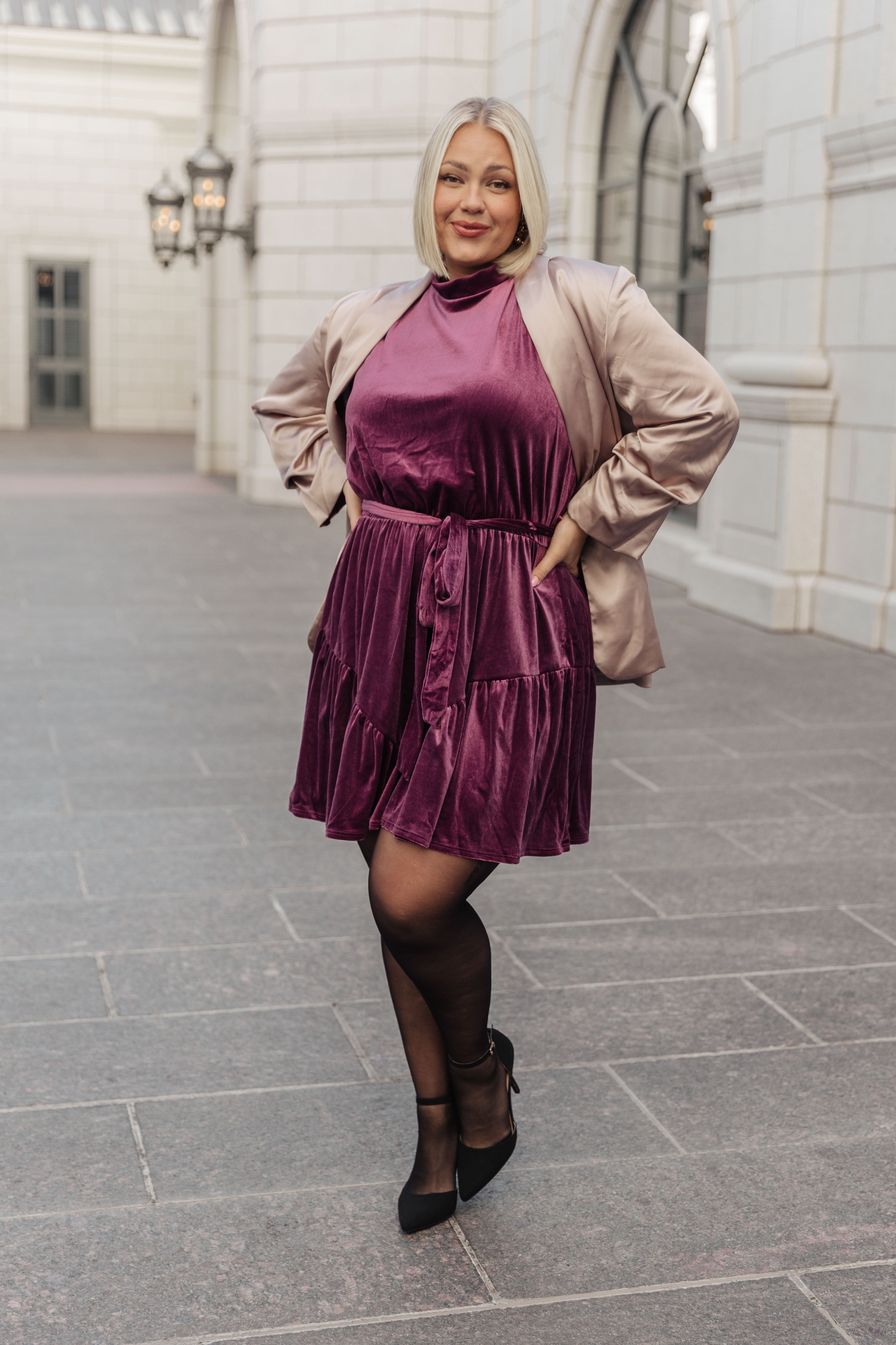 Confident woman wearing a purple velvet bow dress with a beige blazer and black heels outdoors by white building