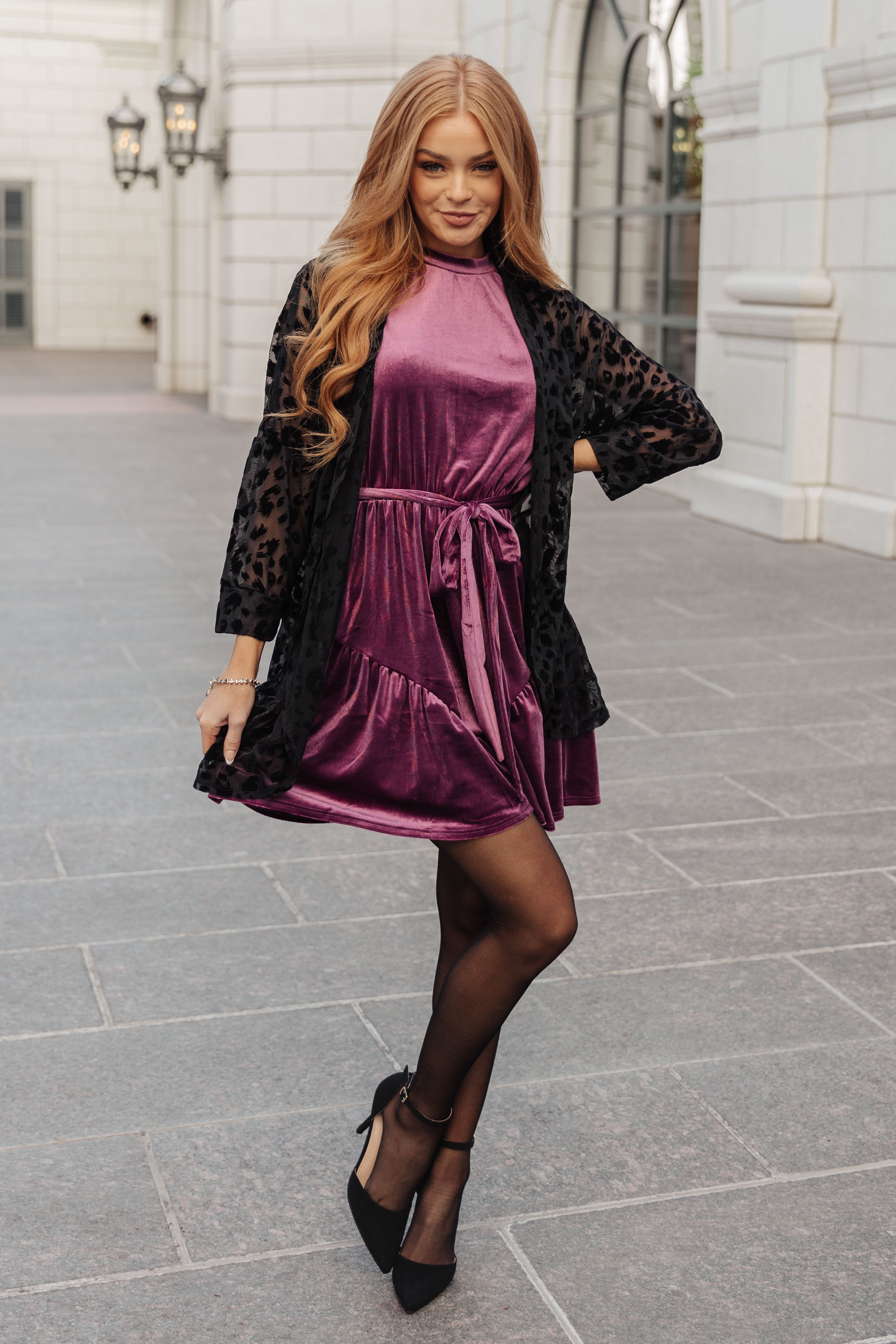 Woman wearing a purple velvet dress tied at the waist with a sheer black leopard-print cardigan outdoors smiling confidently