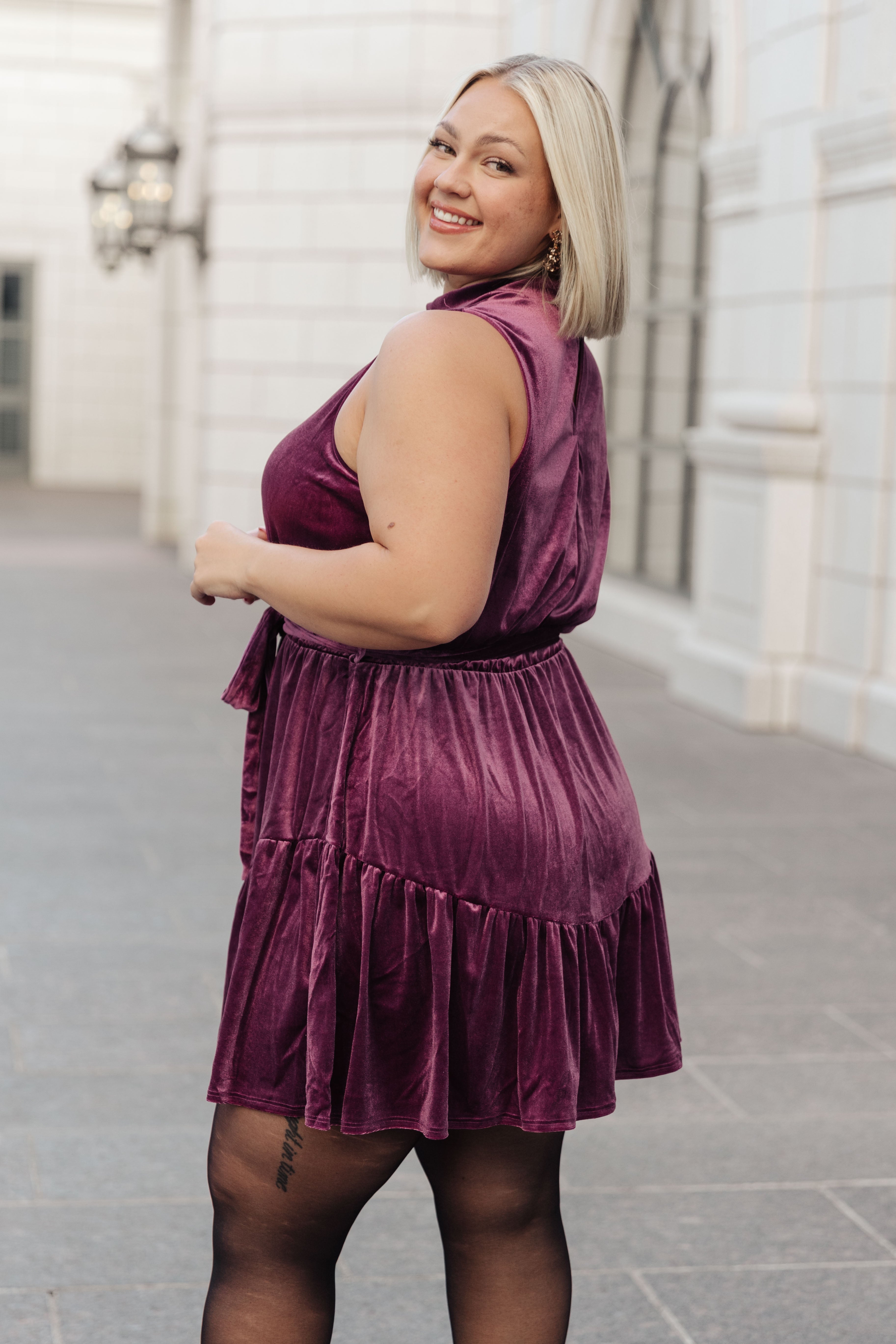 Plus-size woman smiling outdoors wearing a purple velvet dress with a tied waist and ruffled hem paired with black tights