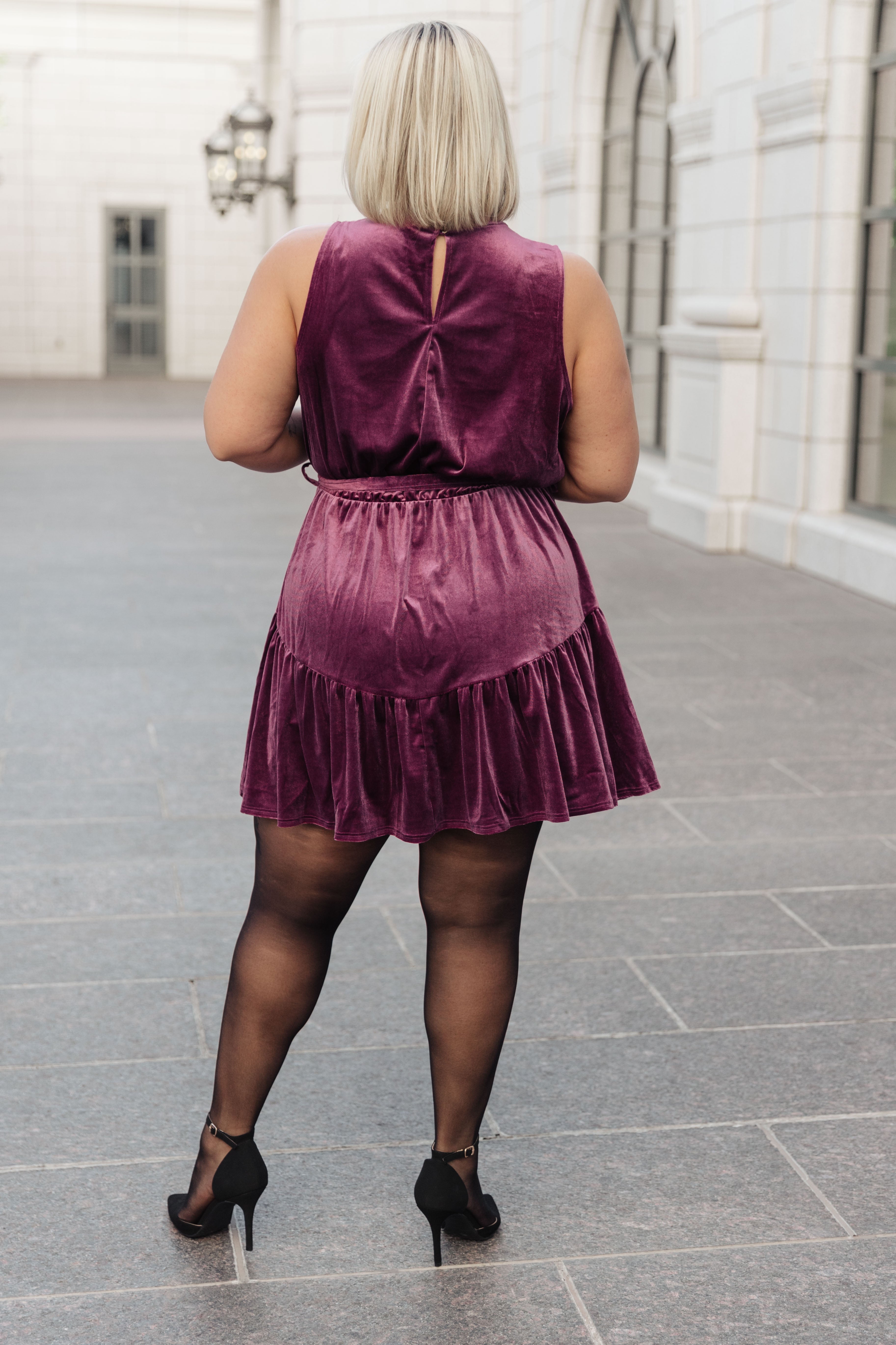 Plus-size woman wearing a purple velvet dress with ruffle hem and sheer black tights, standing outdoors with back facing camera