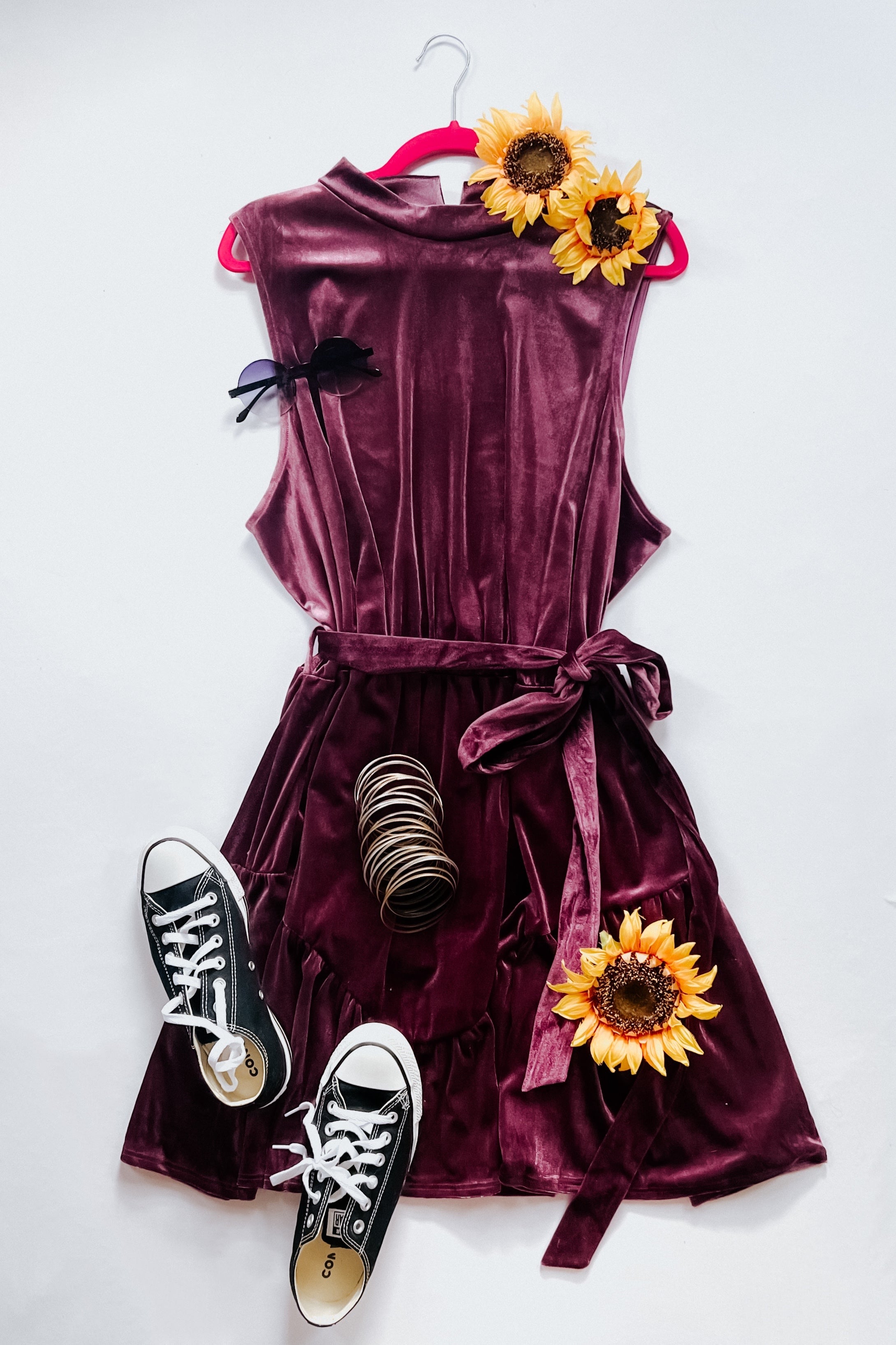 Purple velvet sleeveless dress with tied waist styled with black sneakers, sunglasses, bracelets, and sunflowers on white background