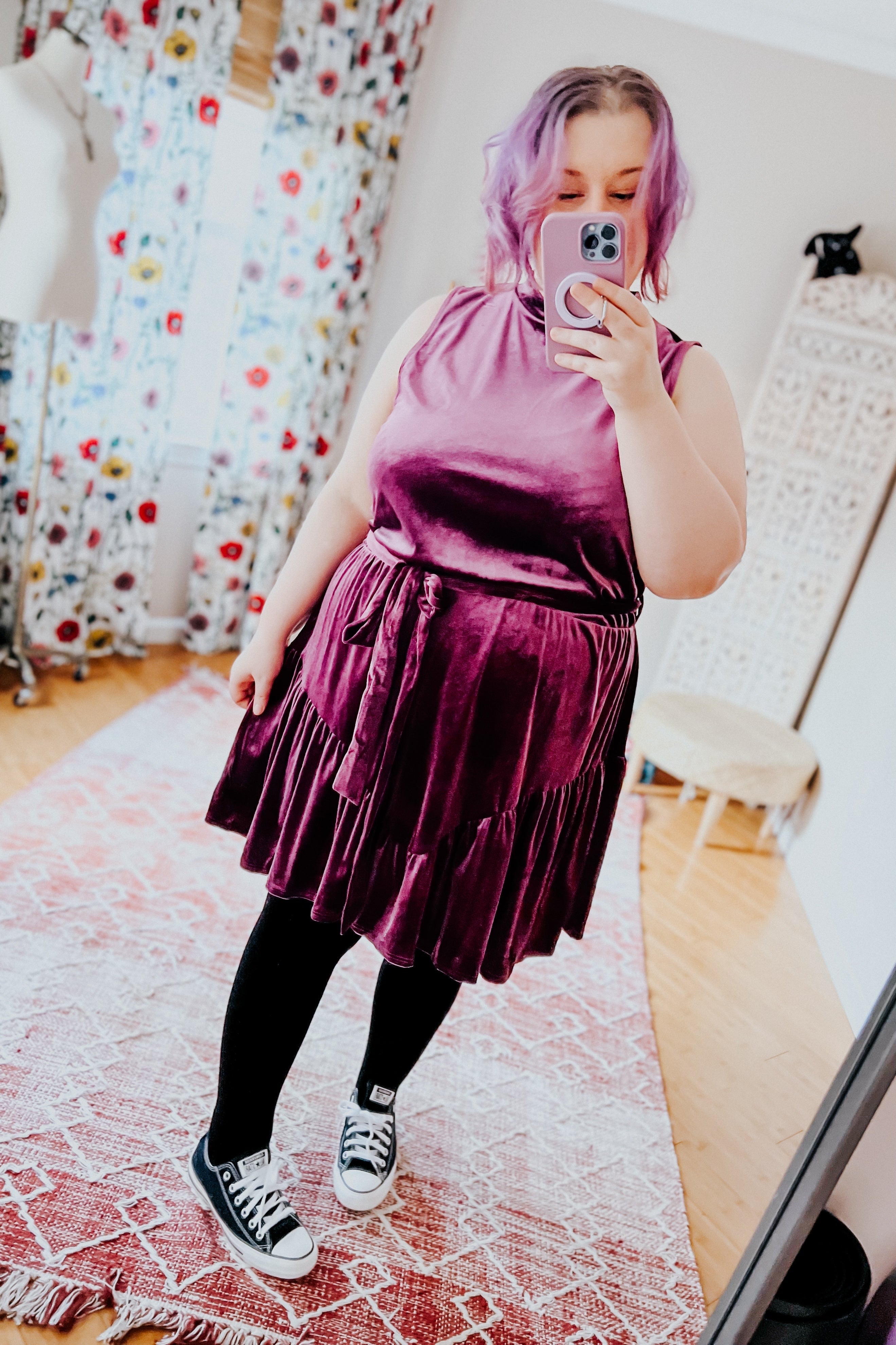 Plus-size woman with purple hair wearing a sleeveless velvet skirt dress and black leggings taking a mirror selfie indoors