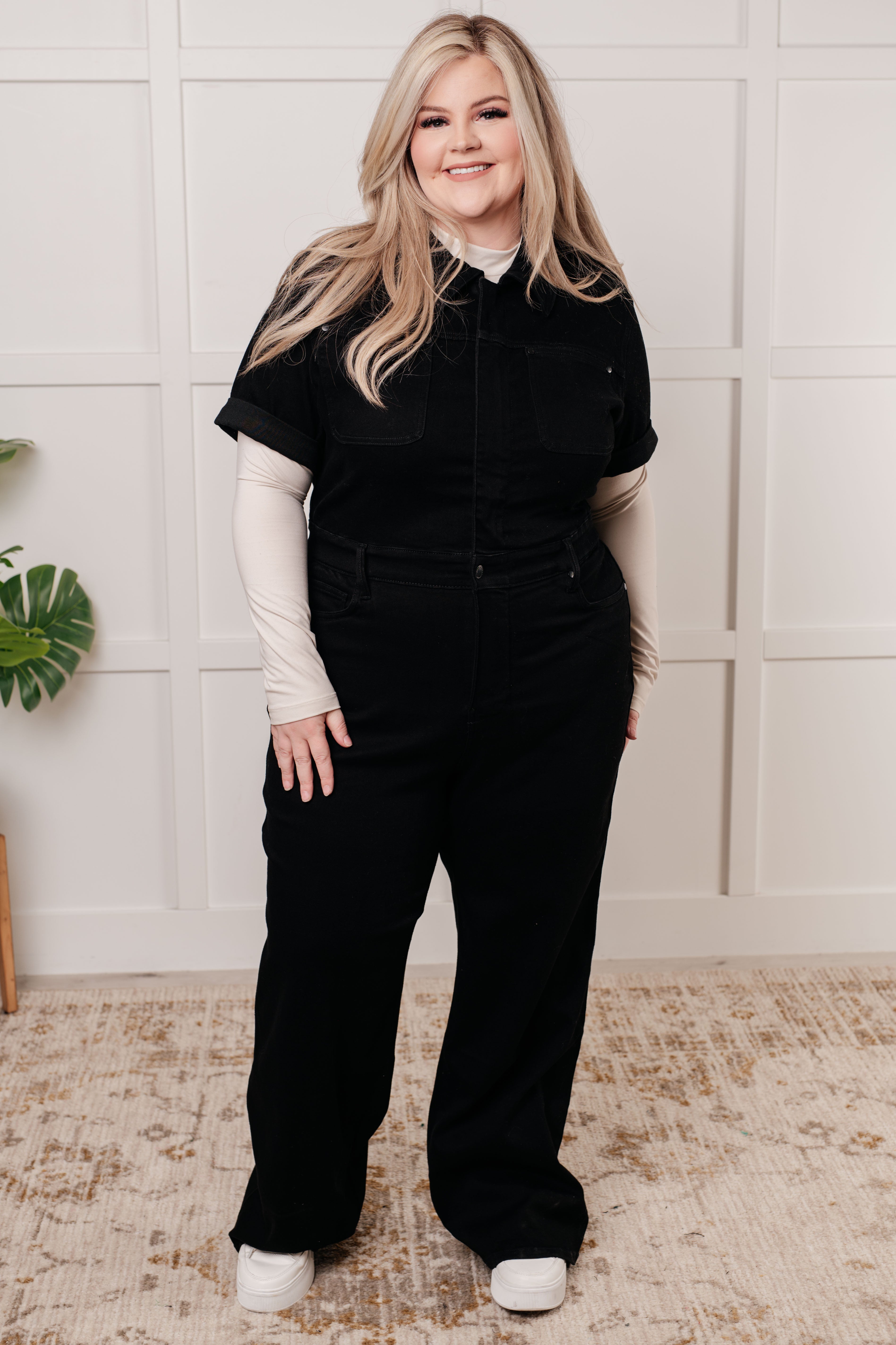 Plus size woman smiling wearing black short sleeve control top denim jumpsuit with beige long sleeve underlayer indoors