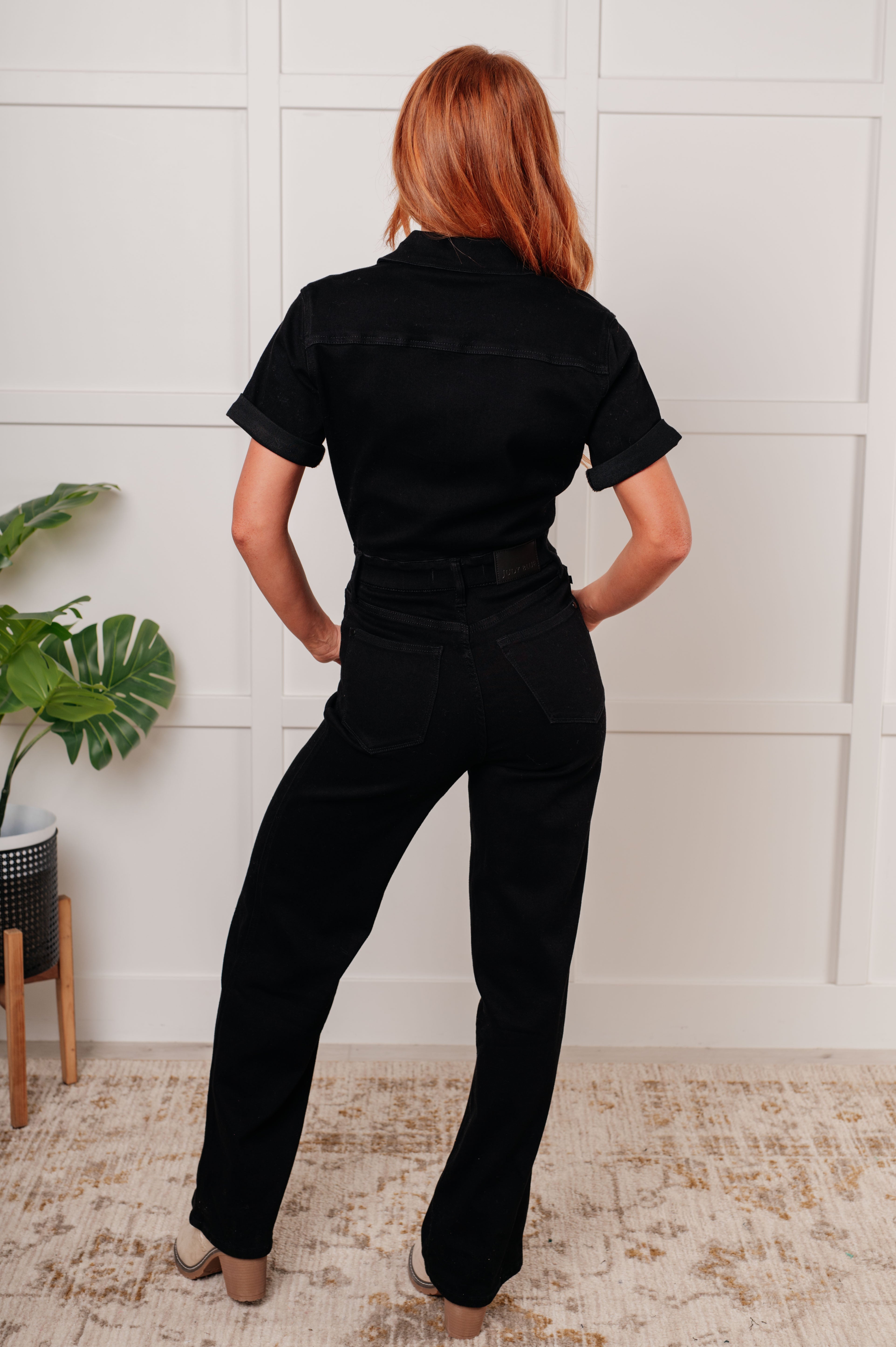 Back view of woman wearing black short sleeve control top denim jumpsuit with rolled sleeves indoors