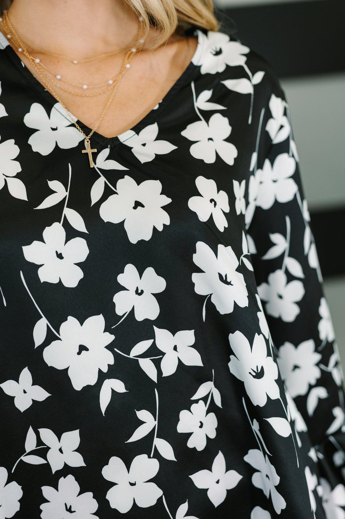 Close up of a woman wearing a black floral blouse with white flowers a V neckline and flowing sleeves with layered gold necklaces