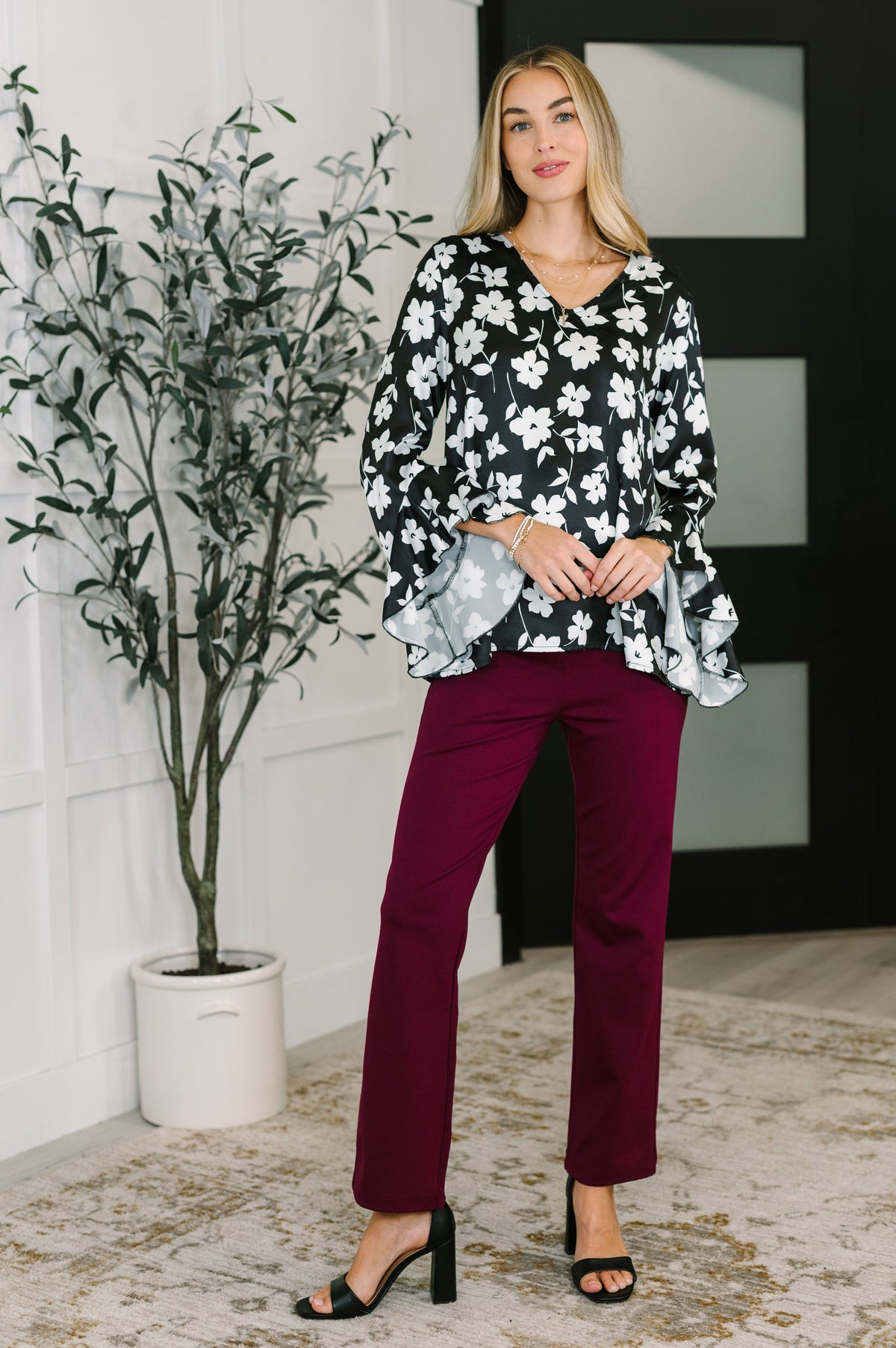 Woman styling a black floral blouse with ruffled sleeves and burgundy pants alongside black heeled sandals in a modern room