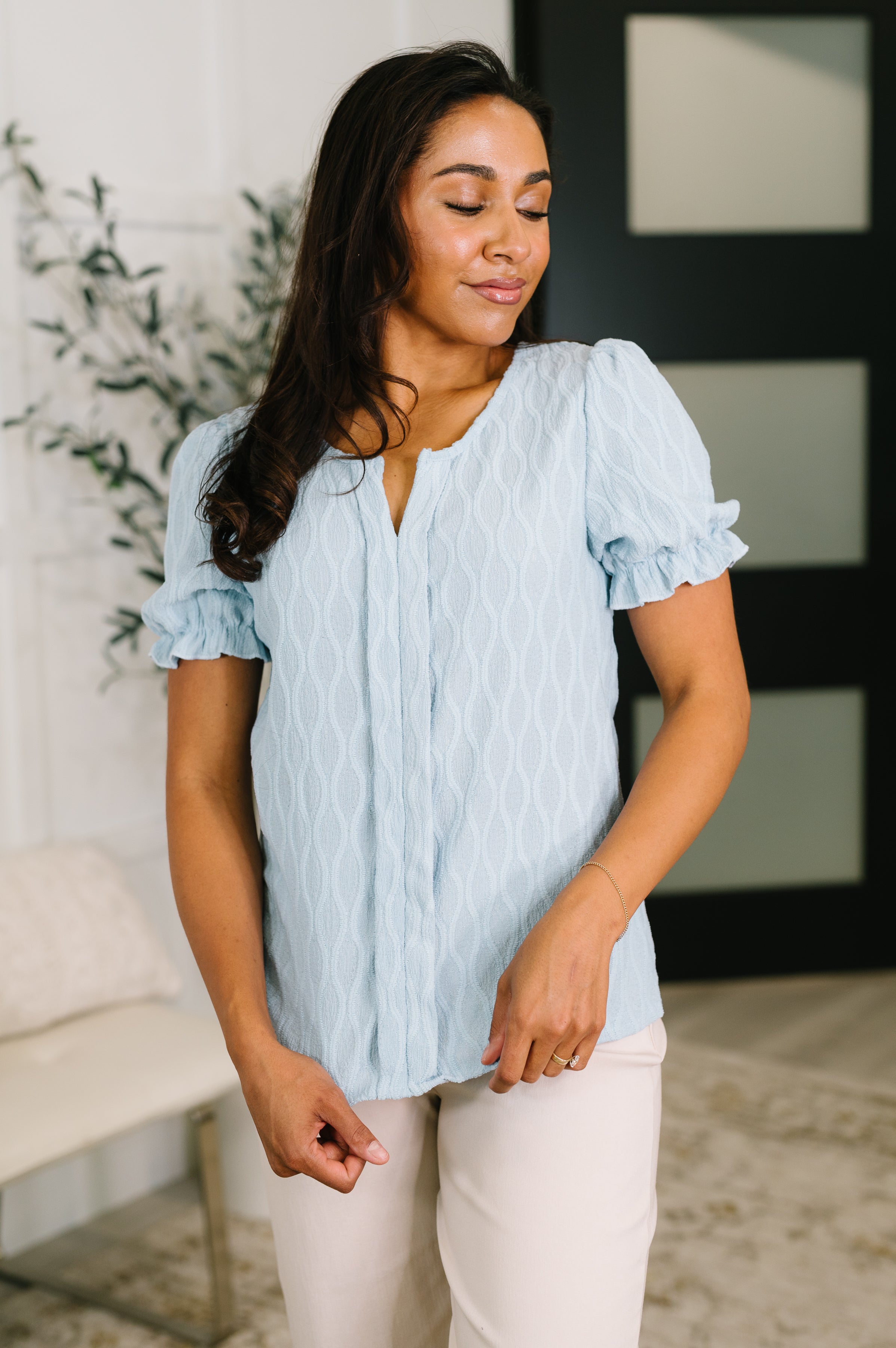 Woman wearing powder blue textured blouse with puffed sleeves standing indoors with eyes closed and slight smile