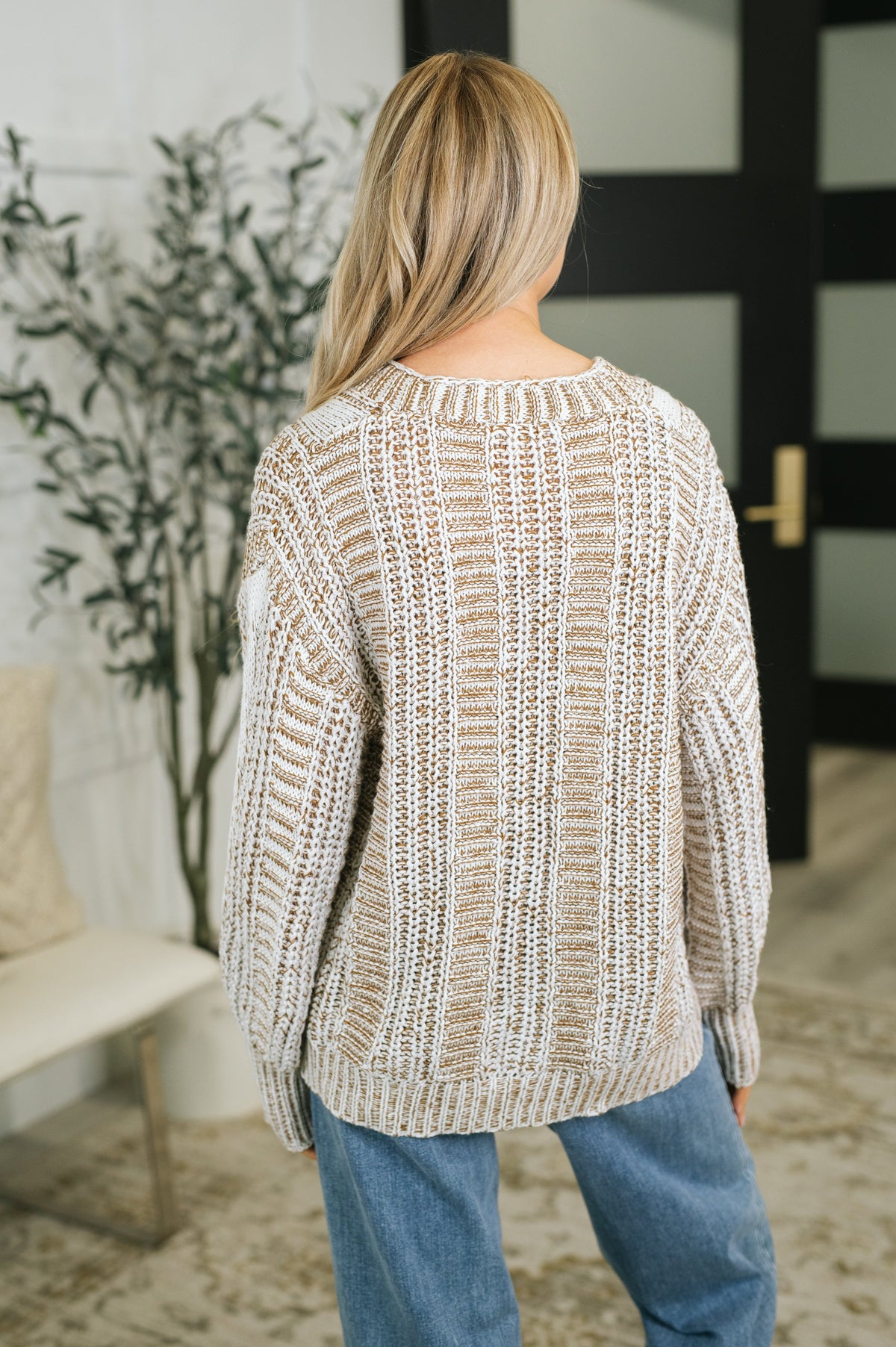 Back view of a woman wearing a beige textured cable knit cardigan with a relaxed fit styled with blue jeans indoors