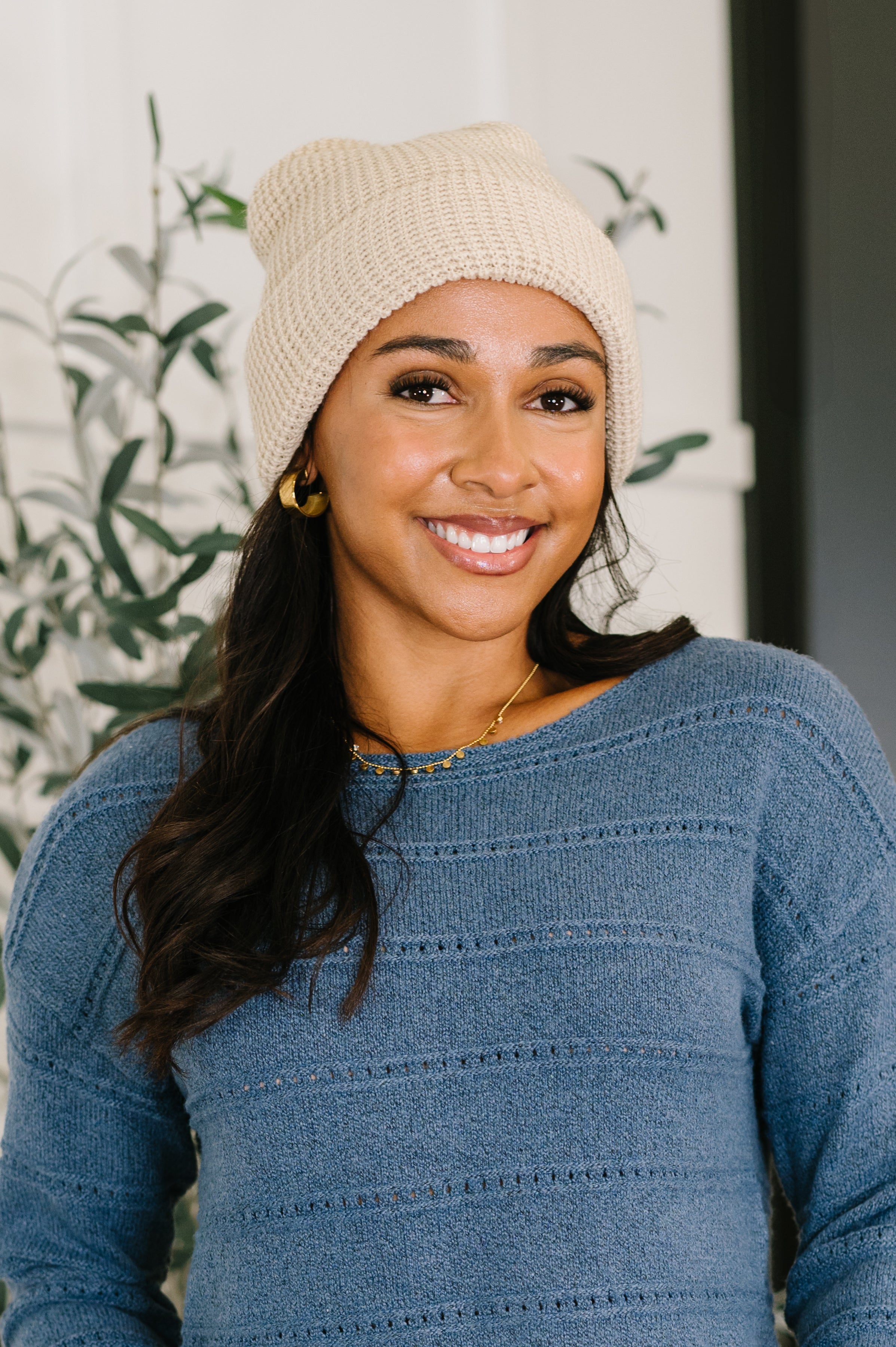 Smiling woman wearing a beige waffle knit beanie with gold hoop earrings and a textured blue sweater indoors