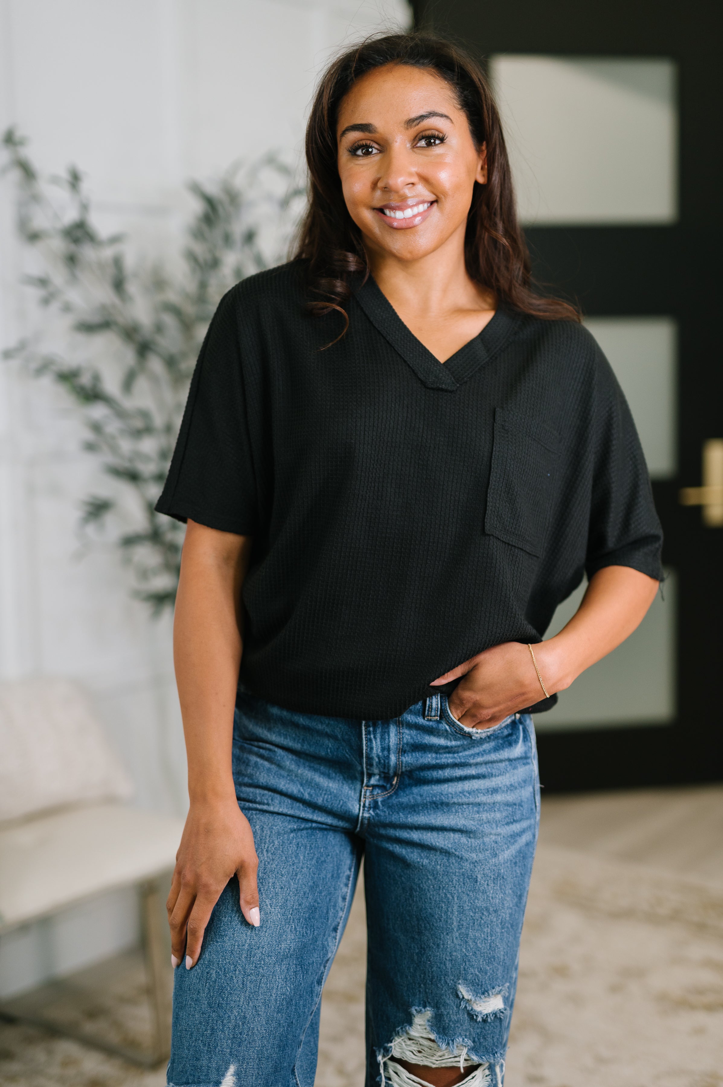 Woman smiling wearing a relaxed black v-neck tee with pocket and distressed blue jeans in a bright, eclectic indoor space