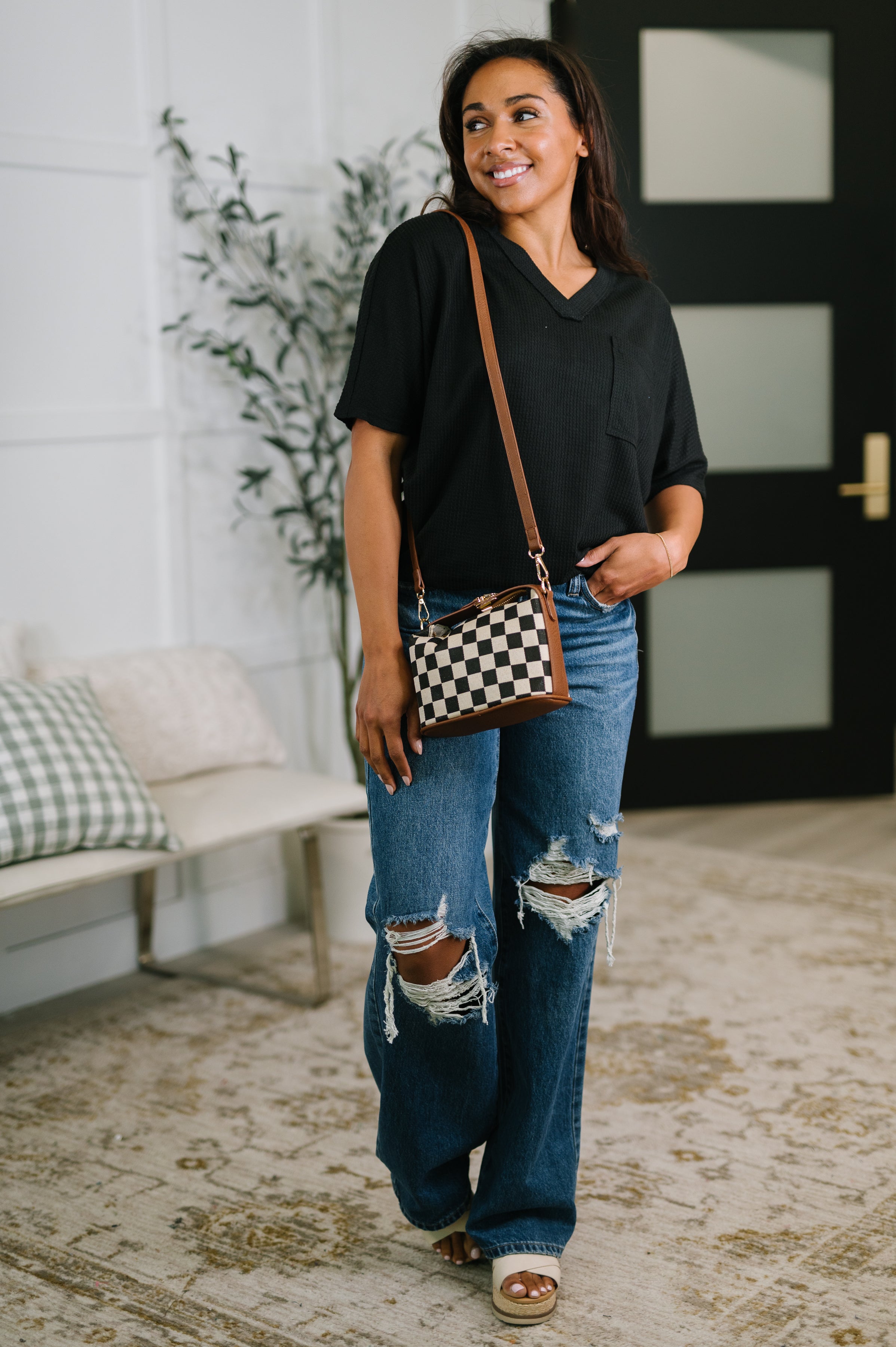 Woman wearing black v-neck tee and ripped jeans holding checkered crossbody bag indoors
