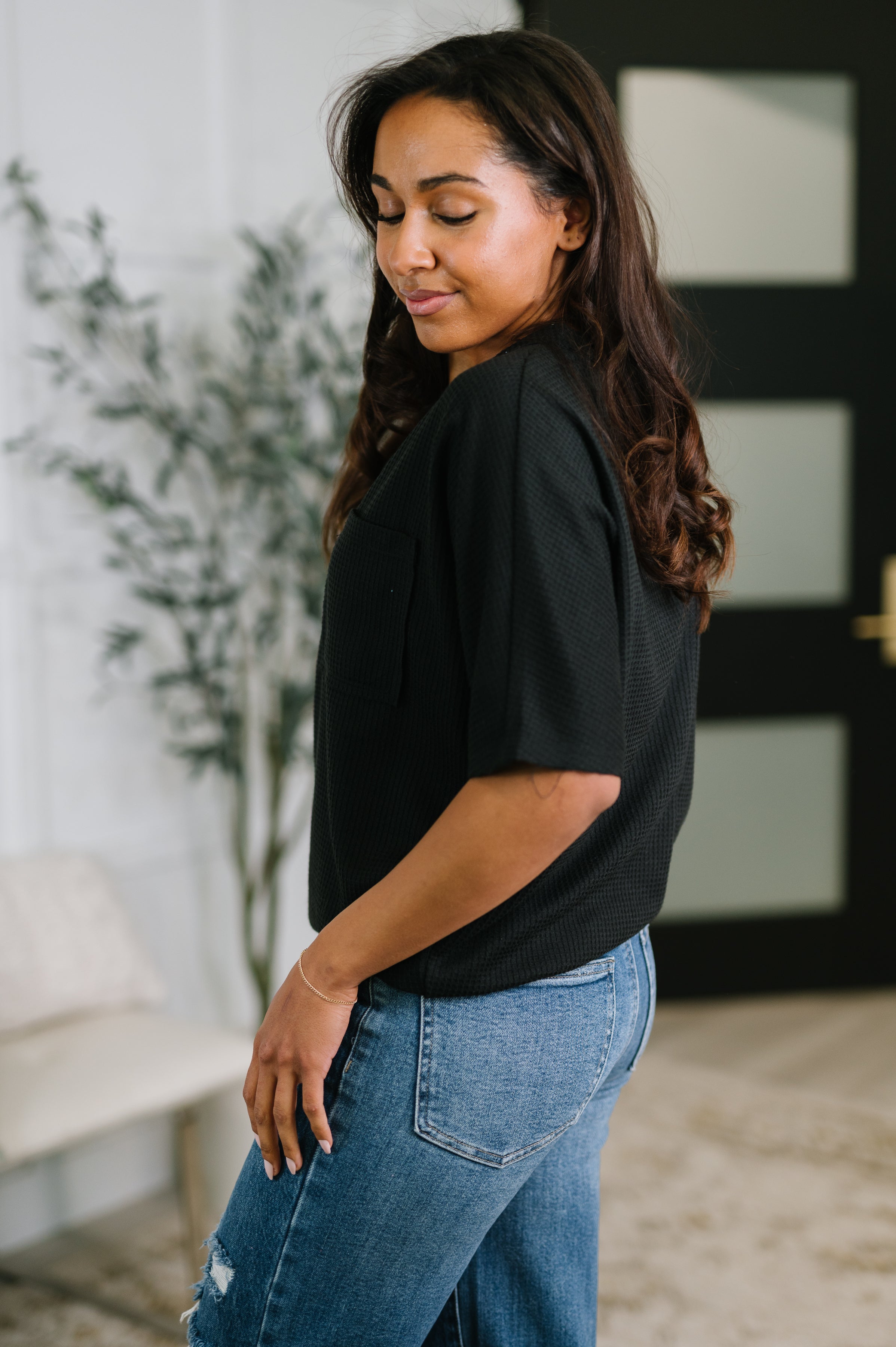 Woman in black loose V-neck top paired with blue distressed jeans standing indoors with eyes closed