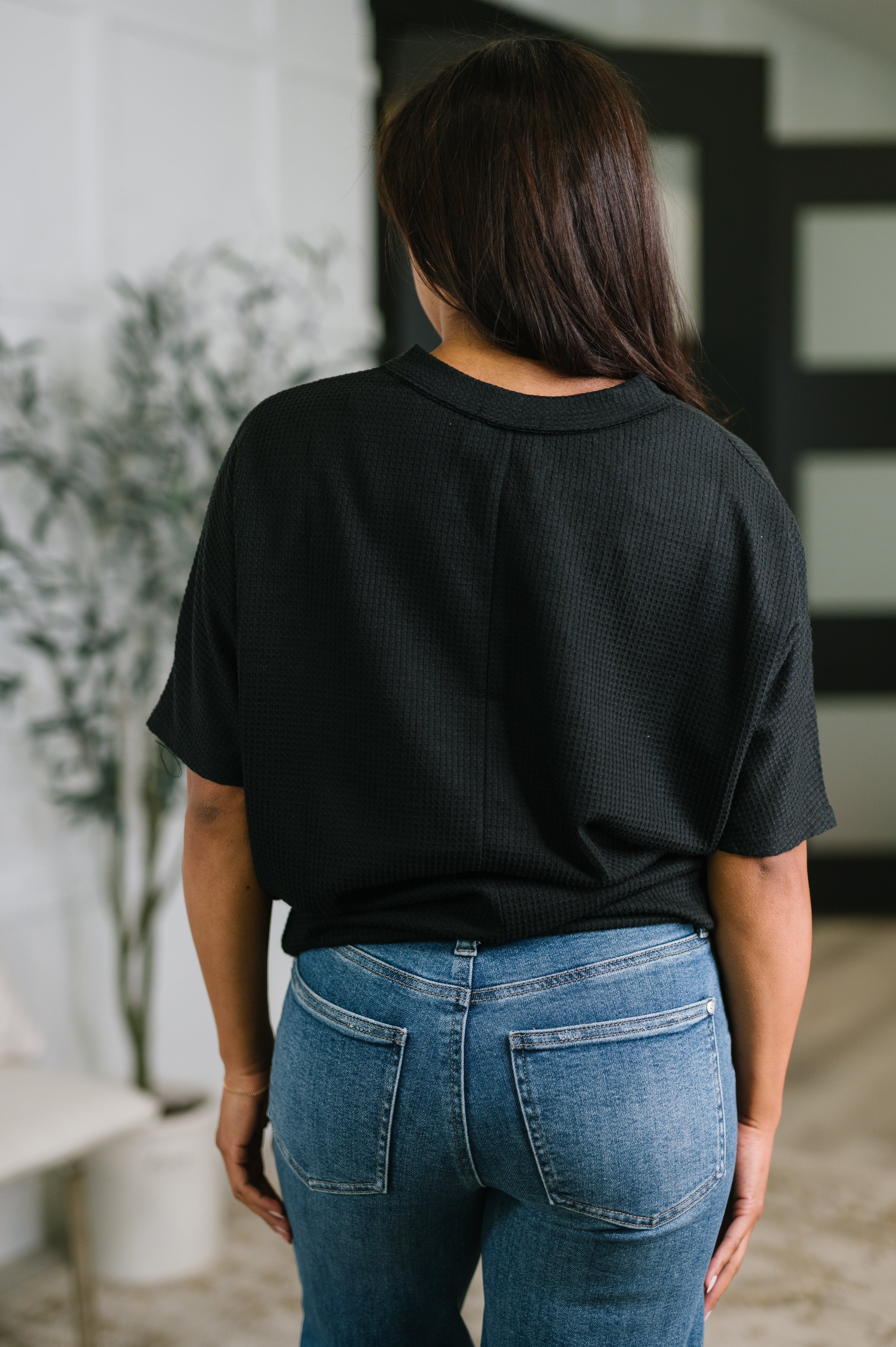 Back view of woman wearing a black textured v-neck tee paired with blue jeans in a relaxed indoor setting