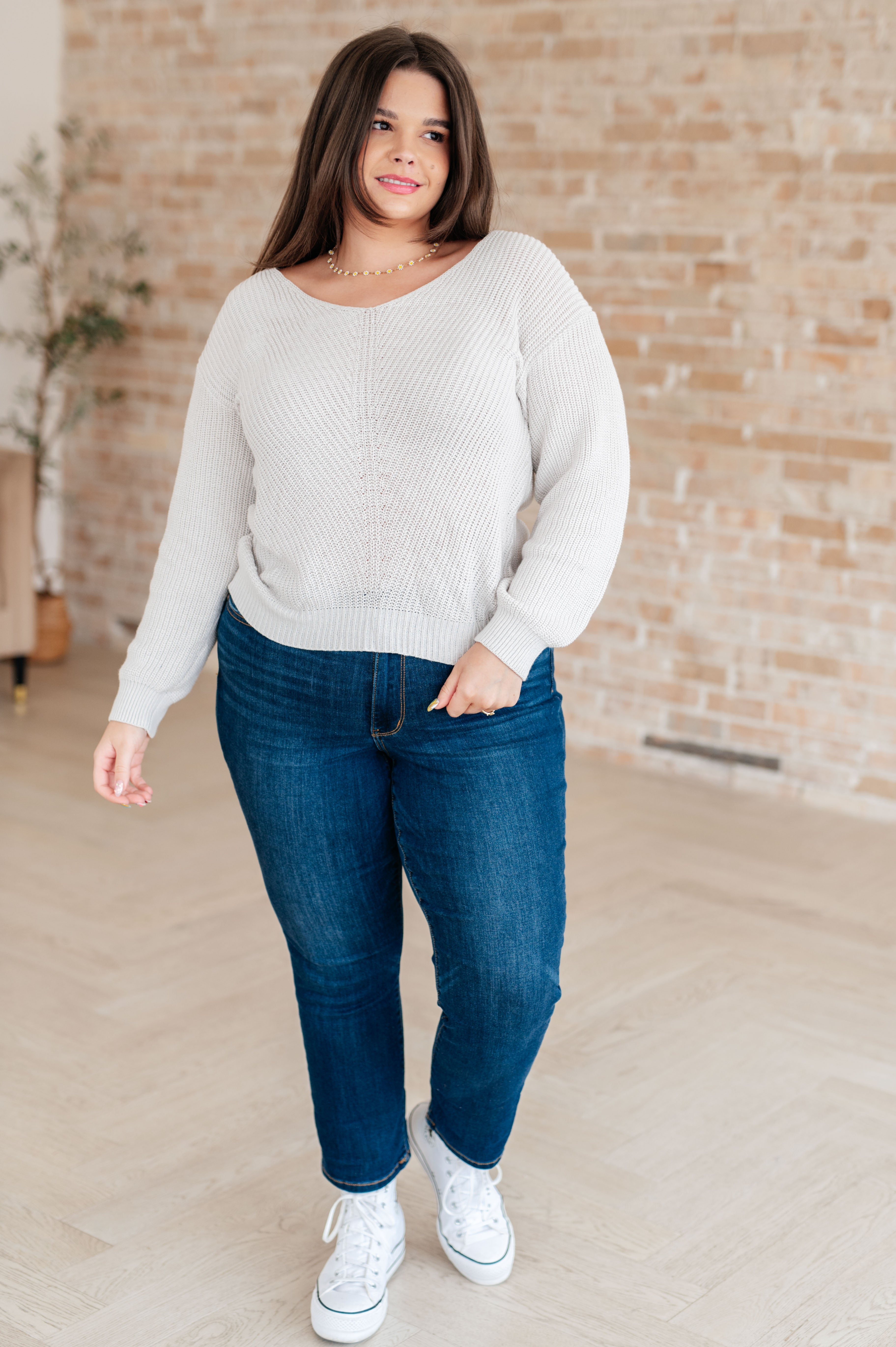 Woman wearing a light grey knit v-neck sweater, blue jeans and white converse sneakers standing against a white brick wall.