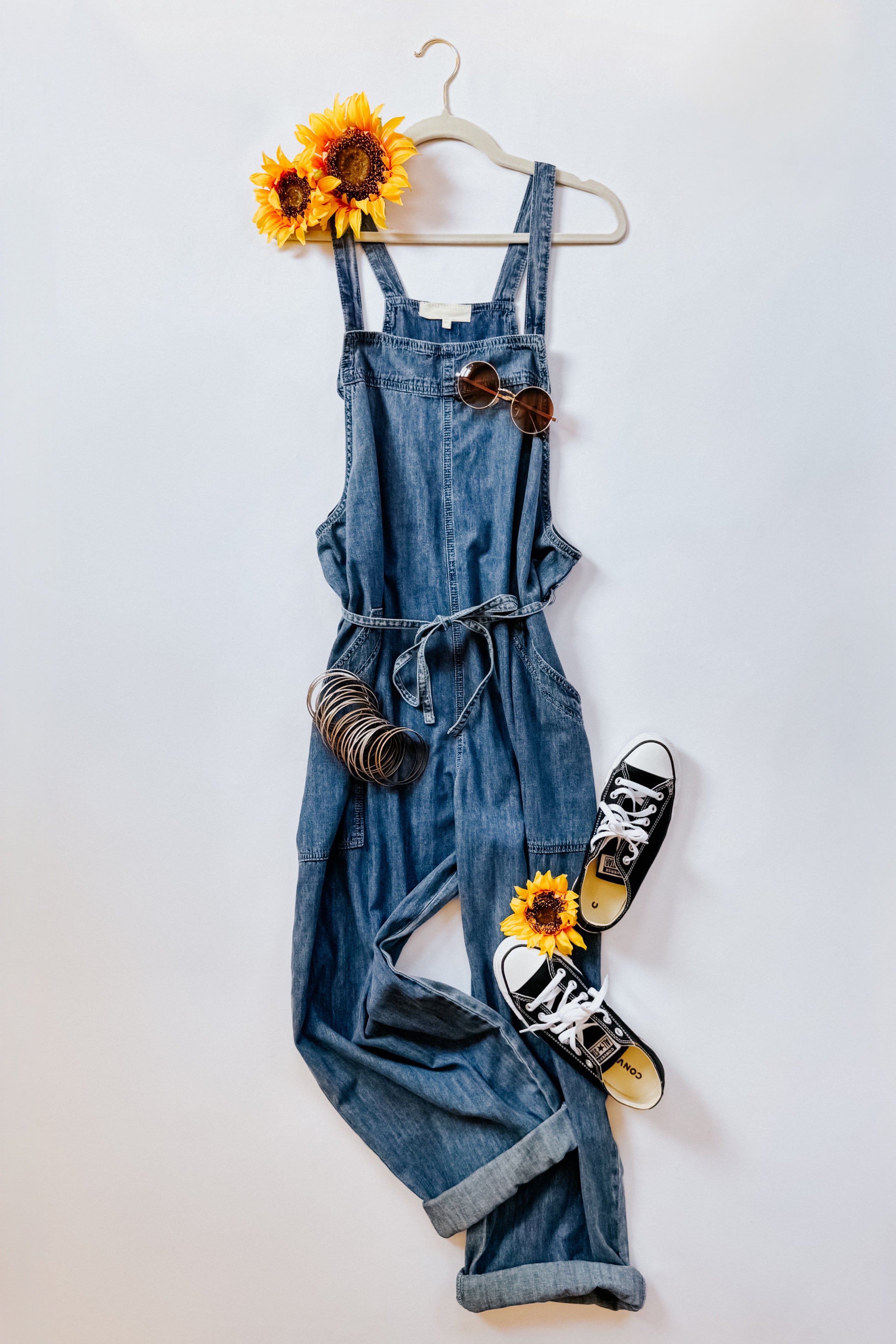 Dark blue denim waist tie overalls styled with sunglasses, bangles, sunflowers, and black canvas sneakers on white background
