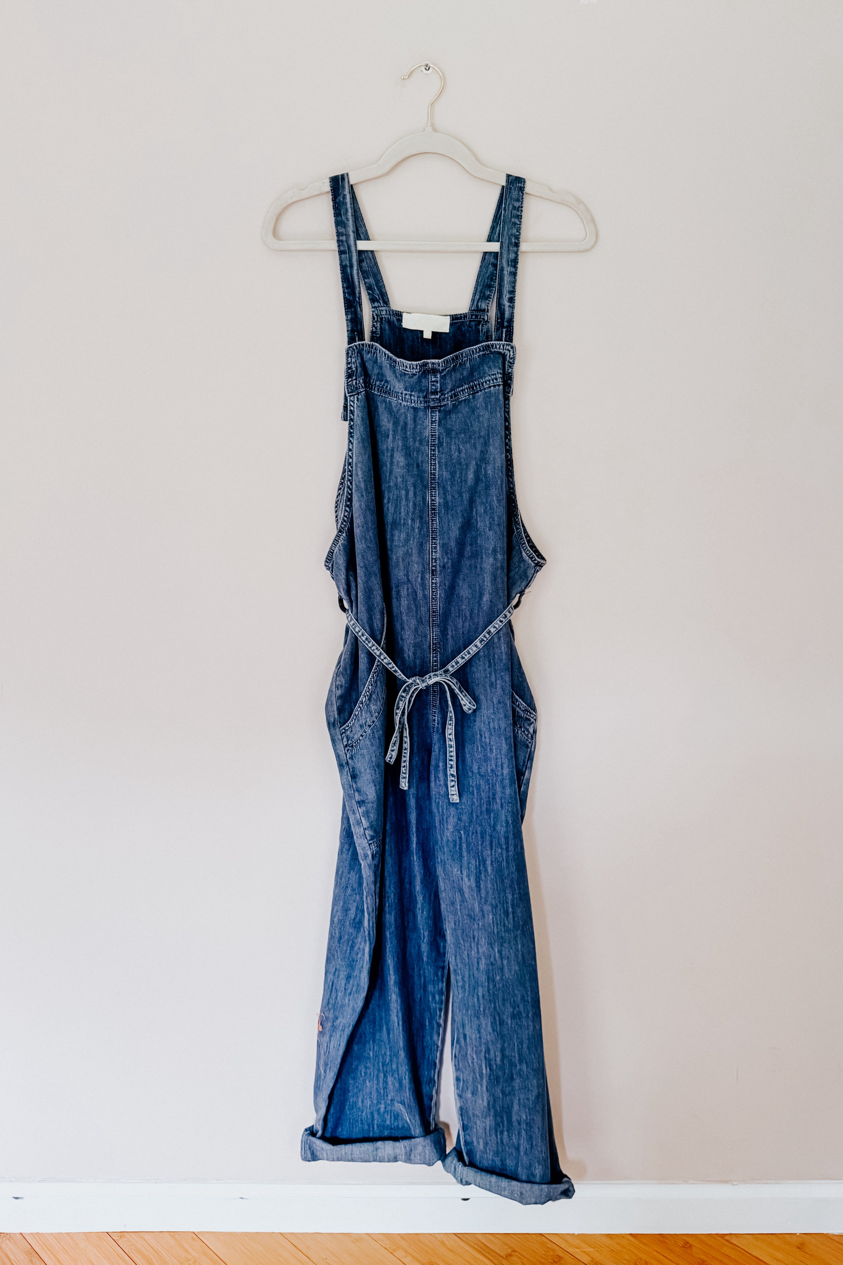 Dark blue denim waist tie overalls hanging on white hanger against light wall background