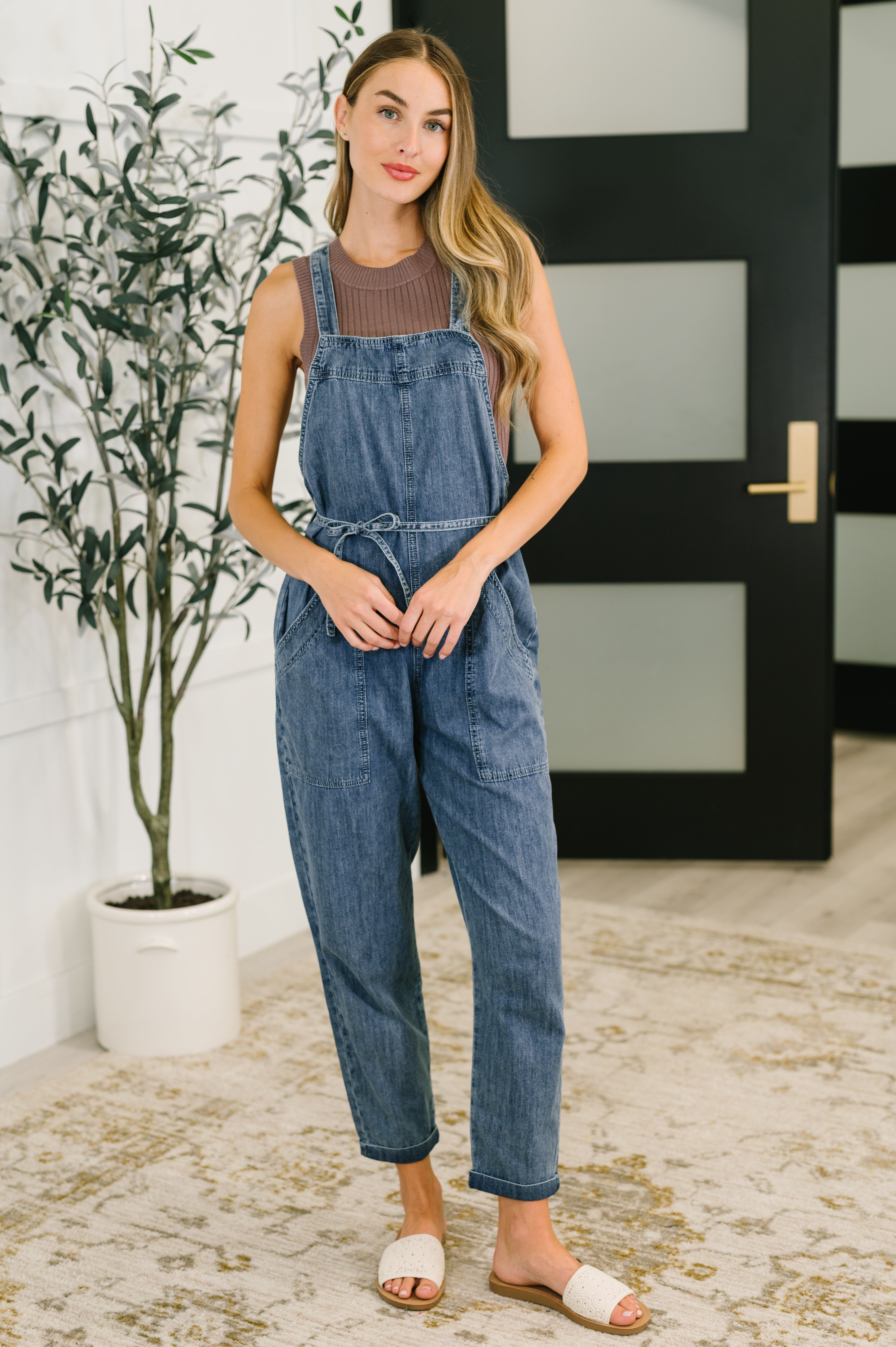 Woman wearing dark blue denim waist tie overalls with sleeveless brown ribbed top and white slide sandals standing indoors