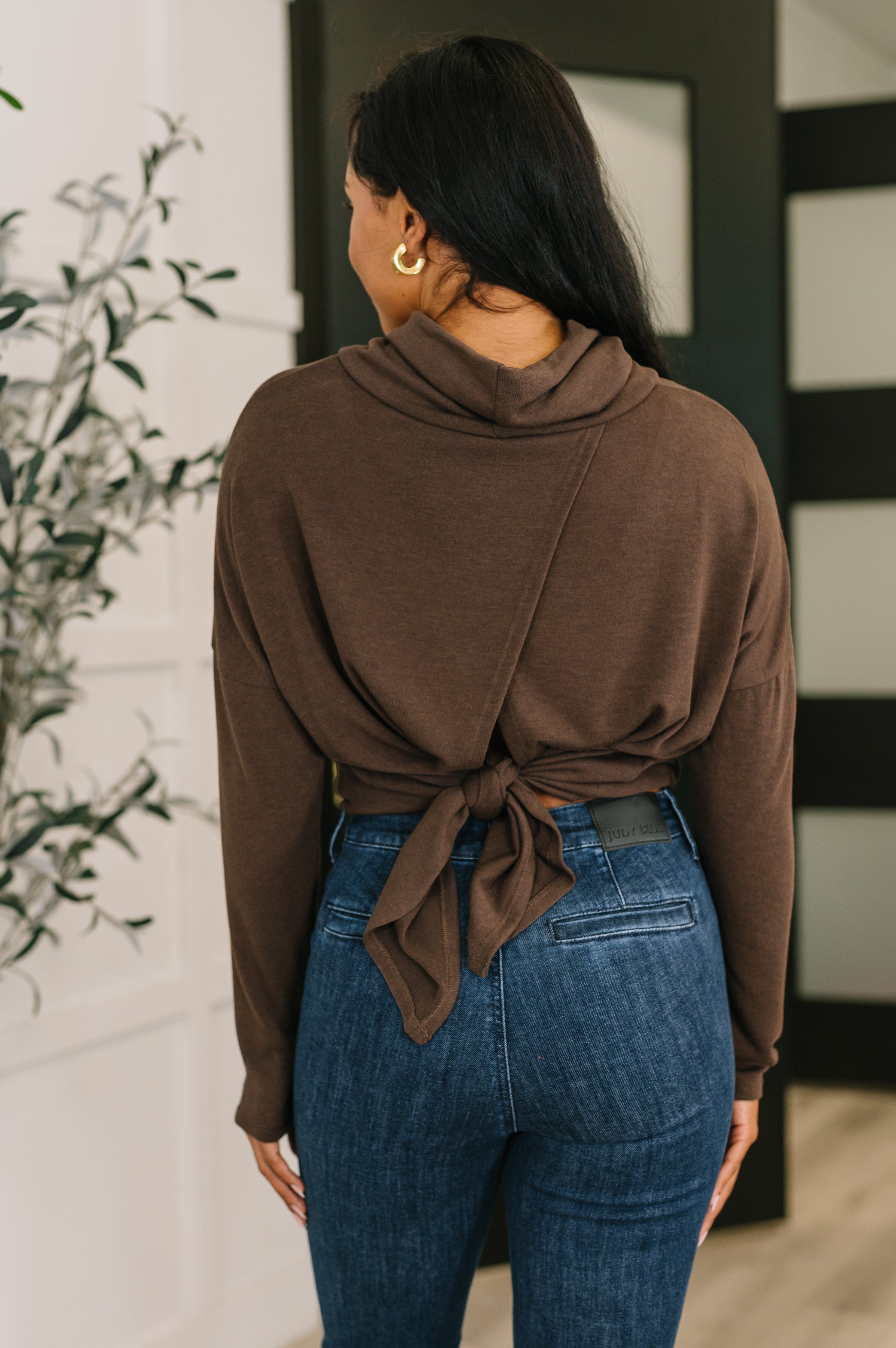 Woman wearing chocolate brown cowl neck pullover tied at the back paired with high-waisted blue jeans indoors