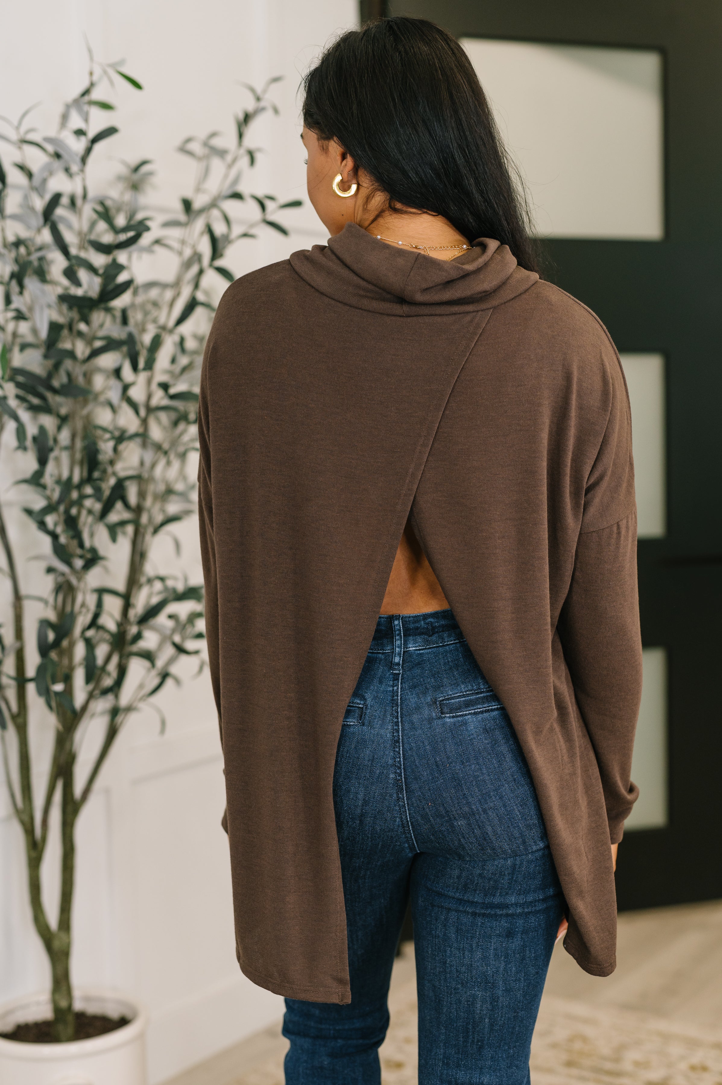 Woman wearing a chocolate brown cowl neck pullover with an open back paired with dark blue jeans indoors
