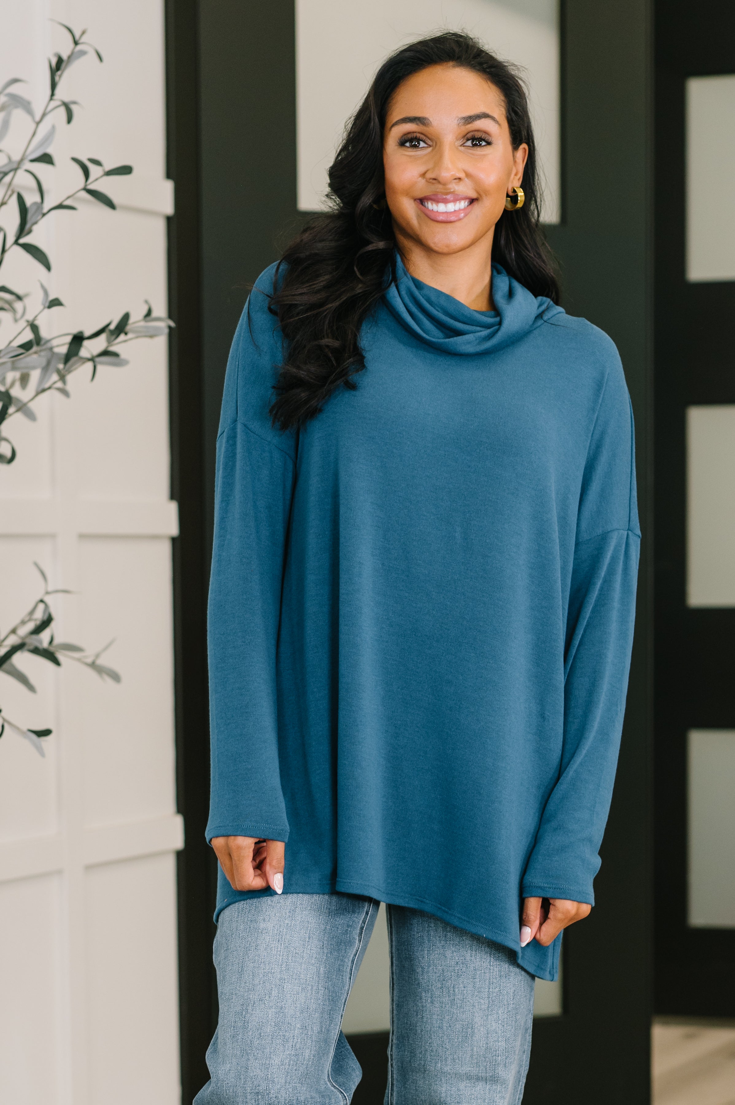Smiling woman wearing a loose dark teal cowl neck pullover with long sleeves paired with light blue jeans indoors