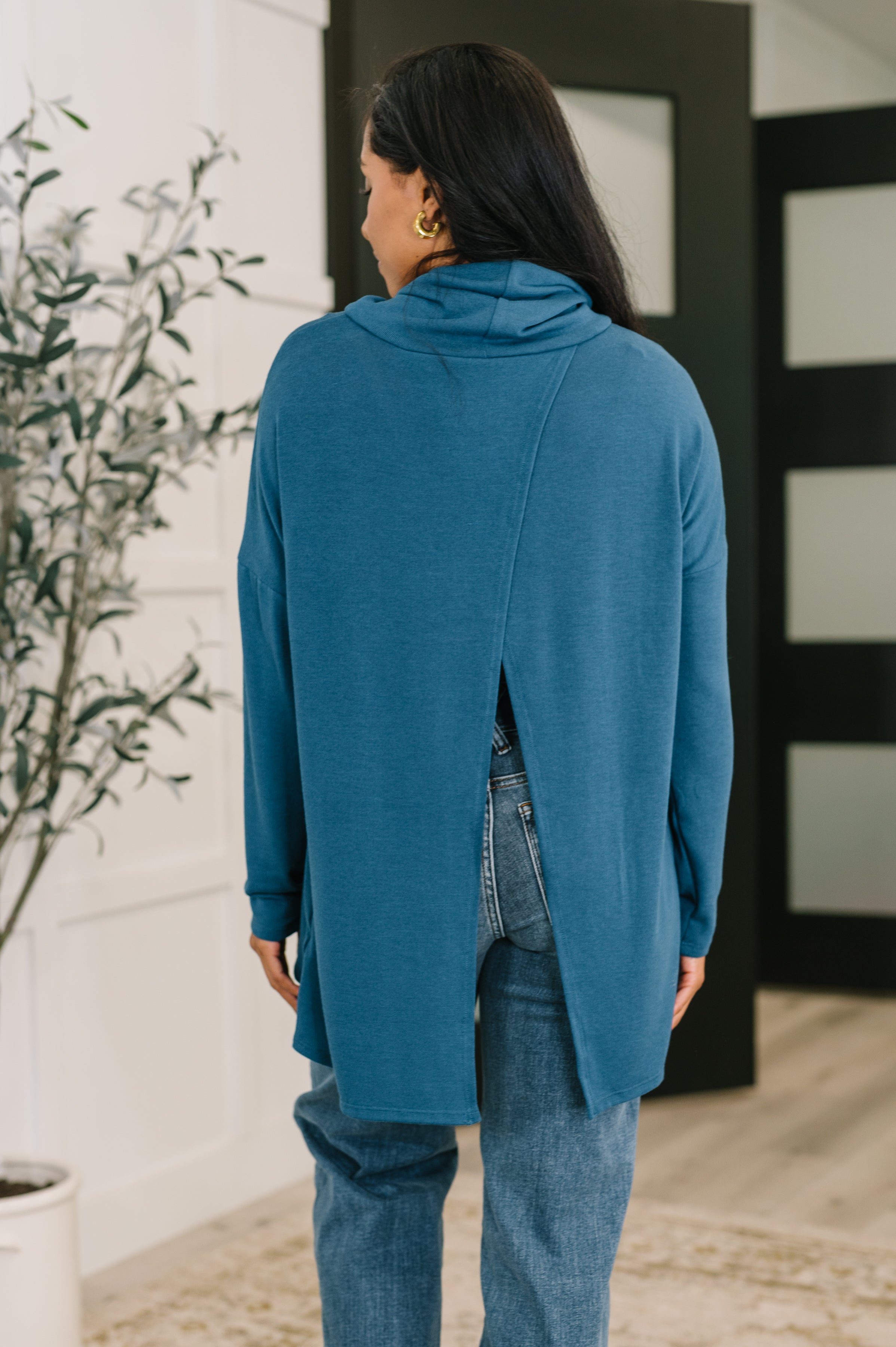 Woman wearing dark teal cowl neck pullover with a back slit paired with blue jeans indoors near plant and door