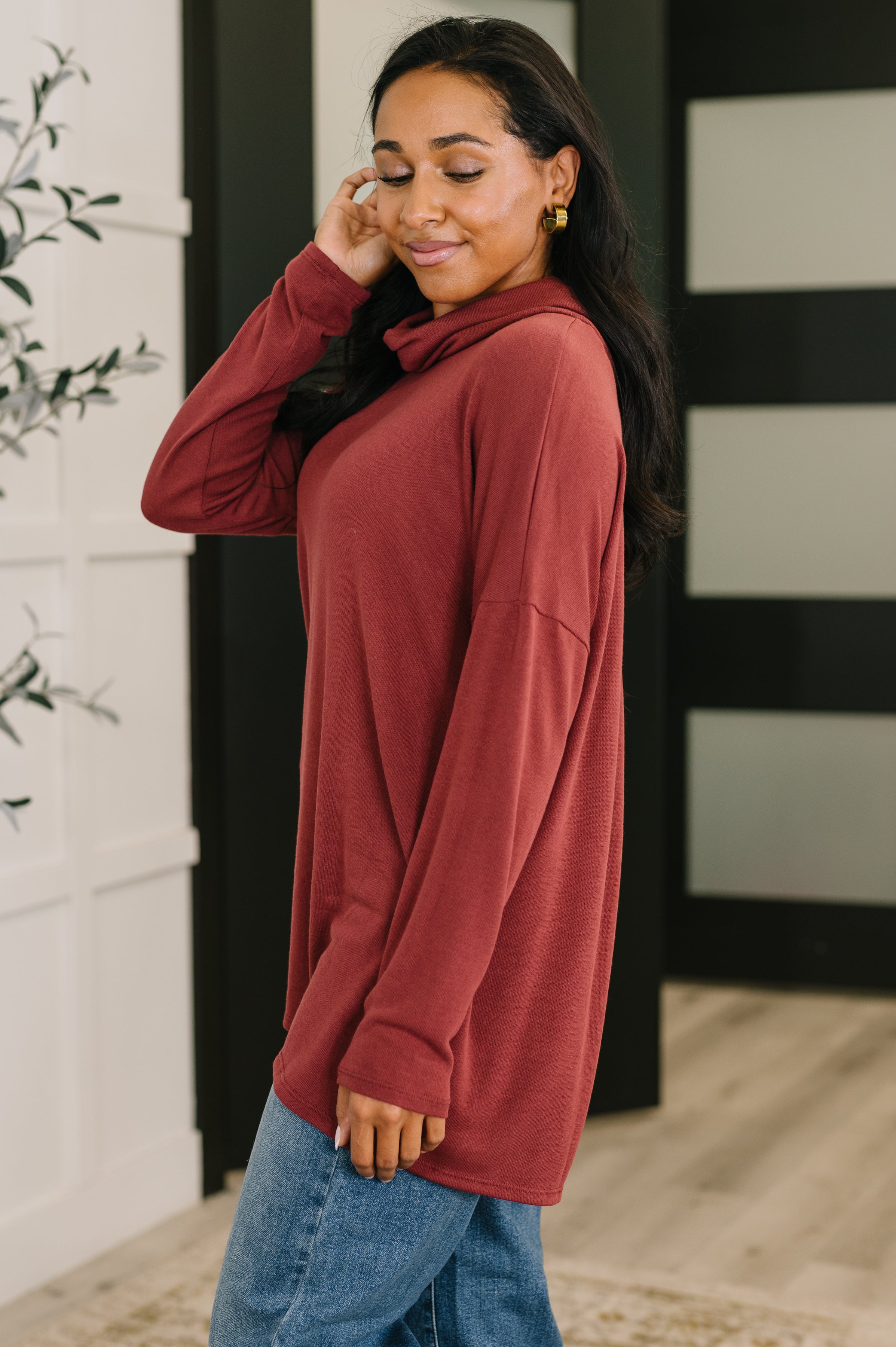 Woman wearing a loose wine-colored cowl neck pullover with long sleeves paired with casual blue jeans indoors