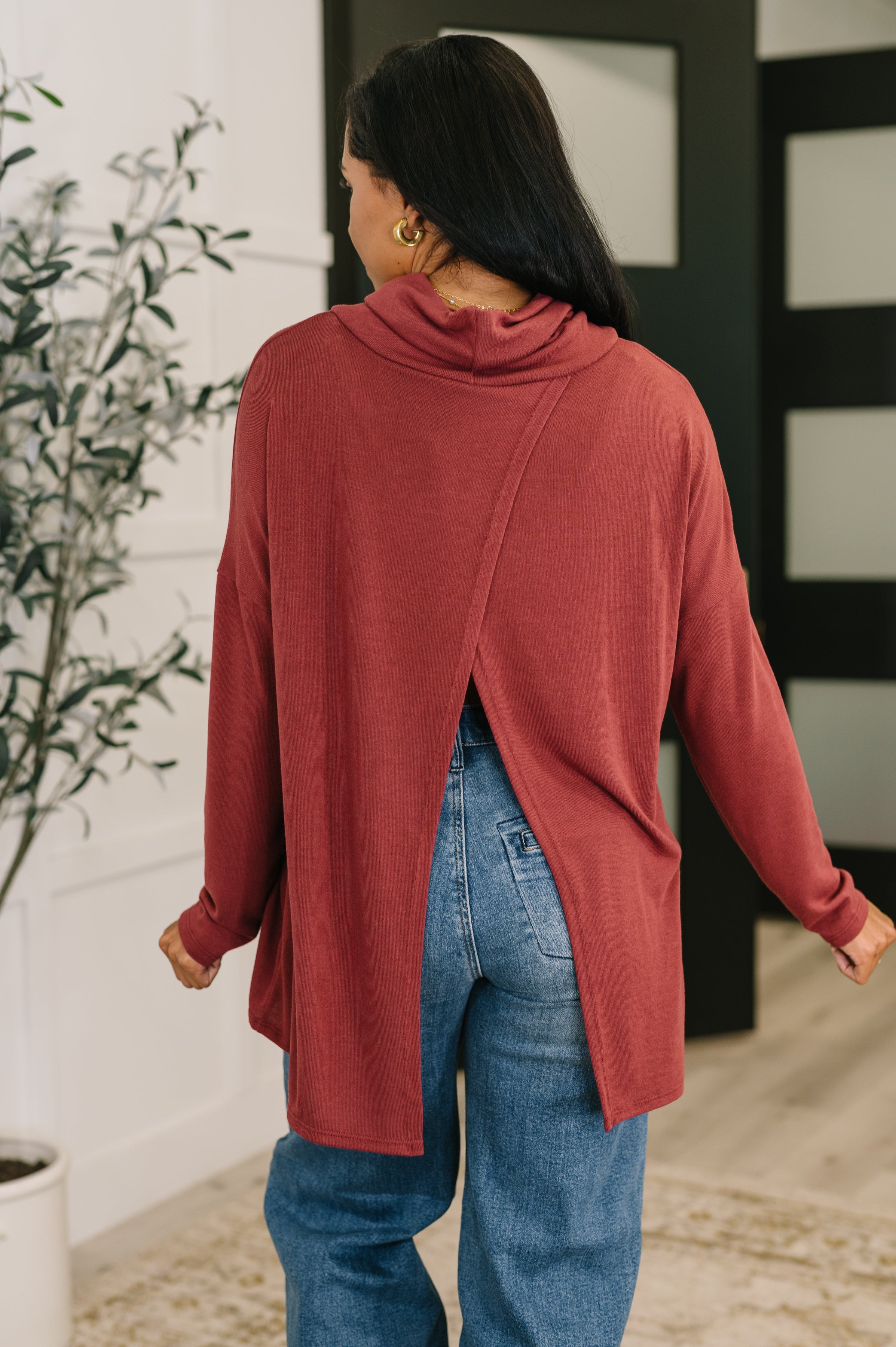 Woman wearing a wine-colored cowl neck pullover with a back slit paired with relaxed blue jeans indoors