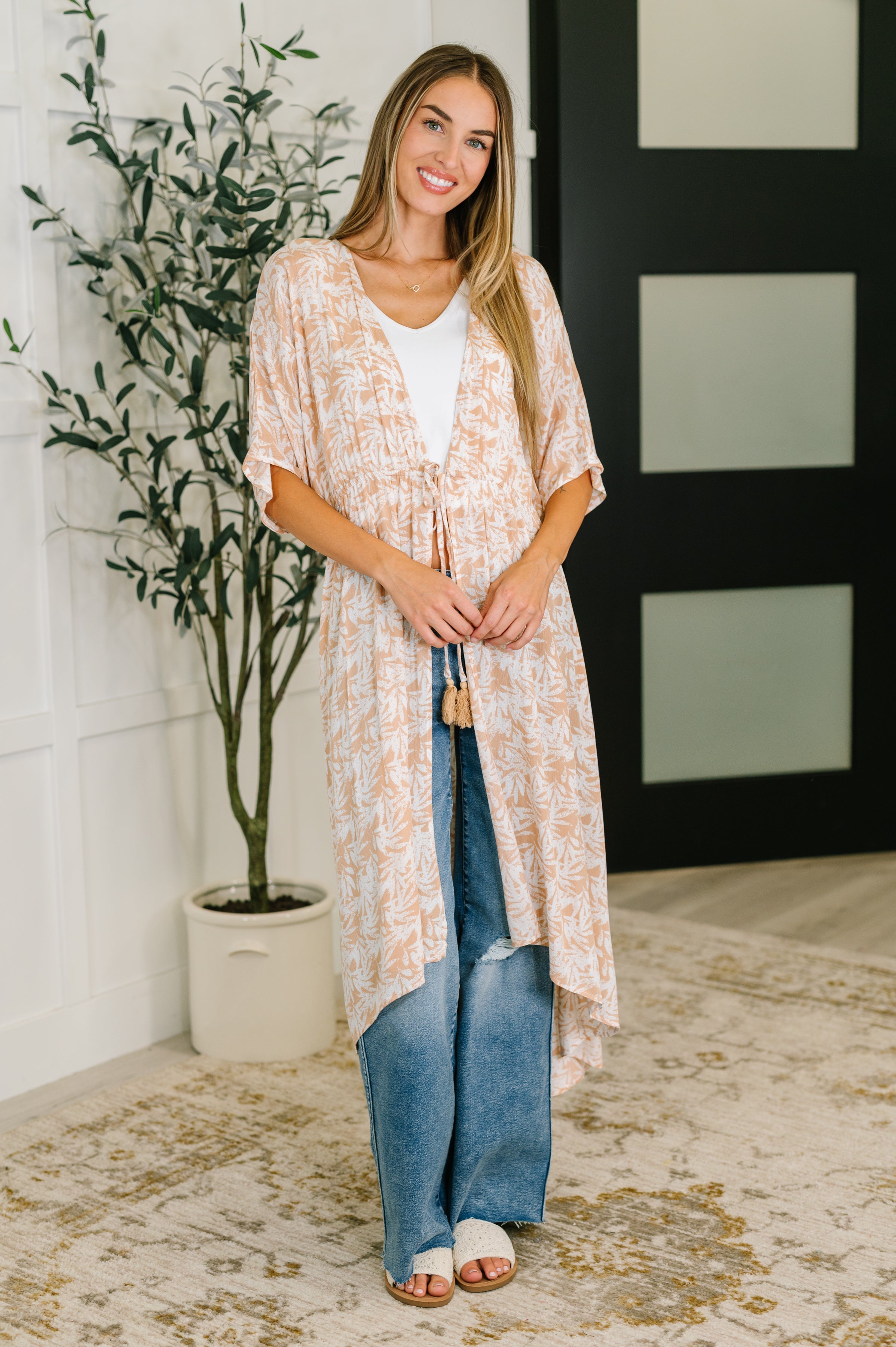 Woman wearing rust tropical print gauze kimono with elastic waist tie over white tank and blue jeans standing indoors