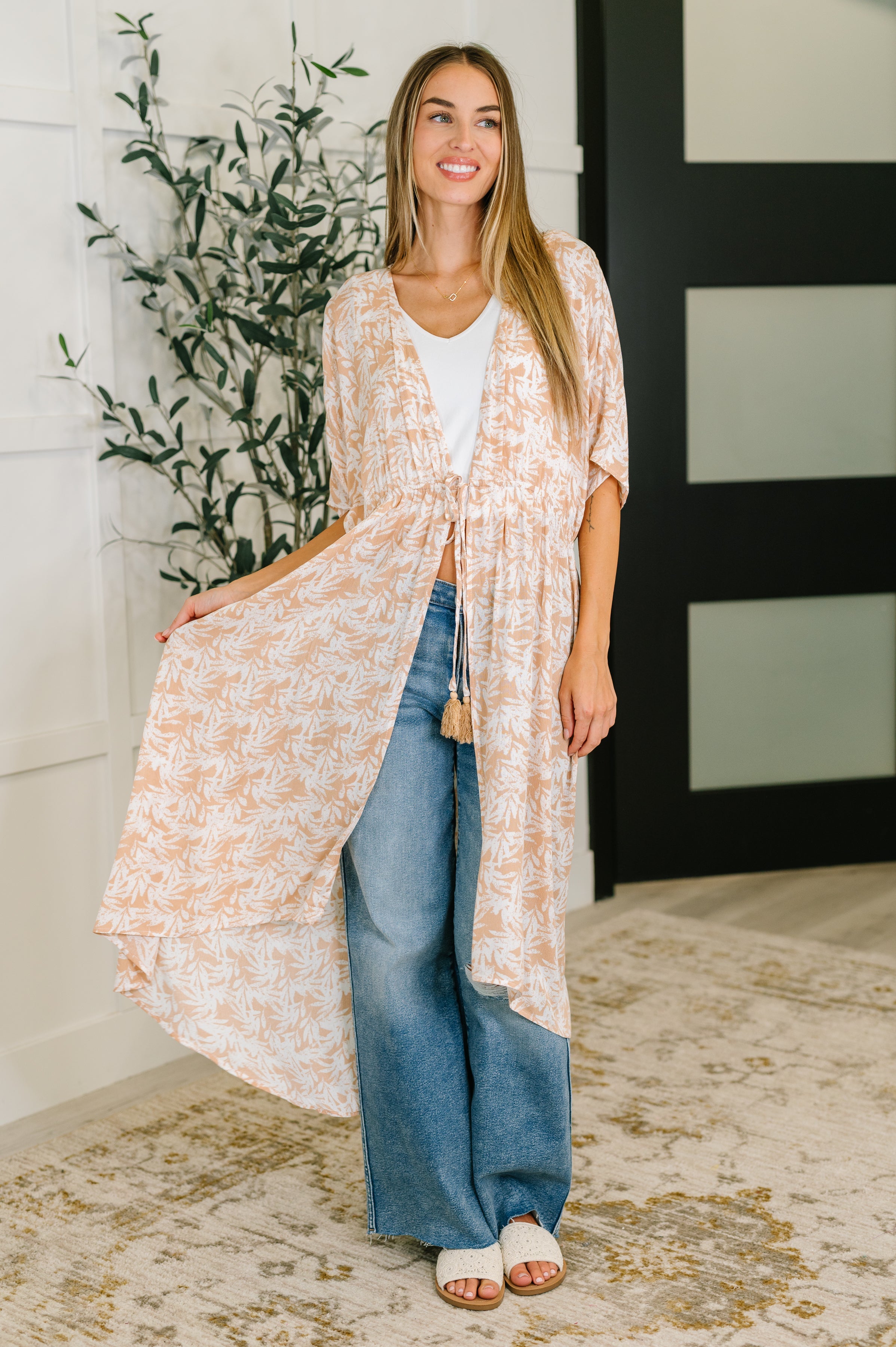 Woman wearing rust tropical print lightweight kimono with elastic waist tie over white top and wide-leg jeans indoors
