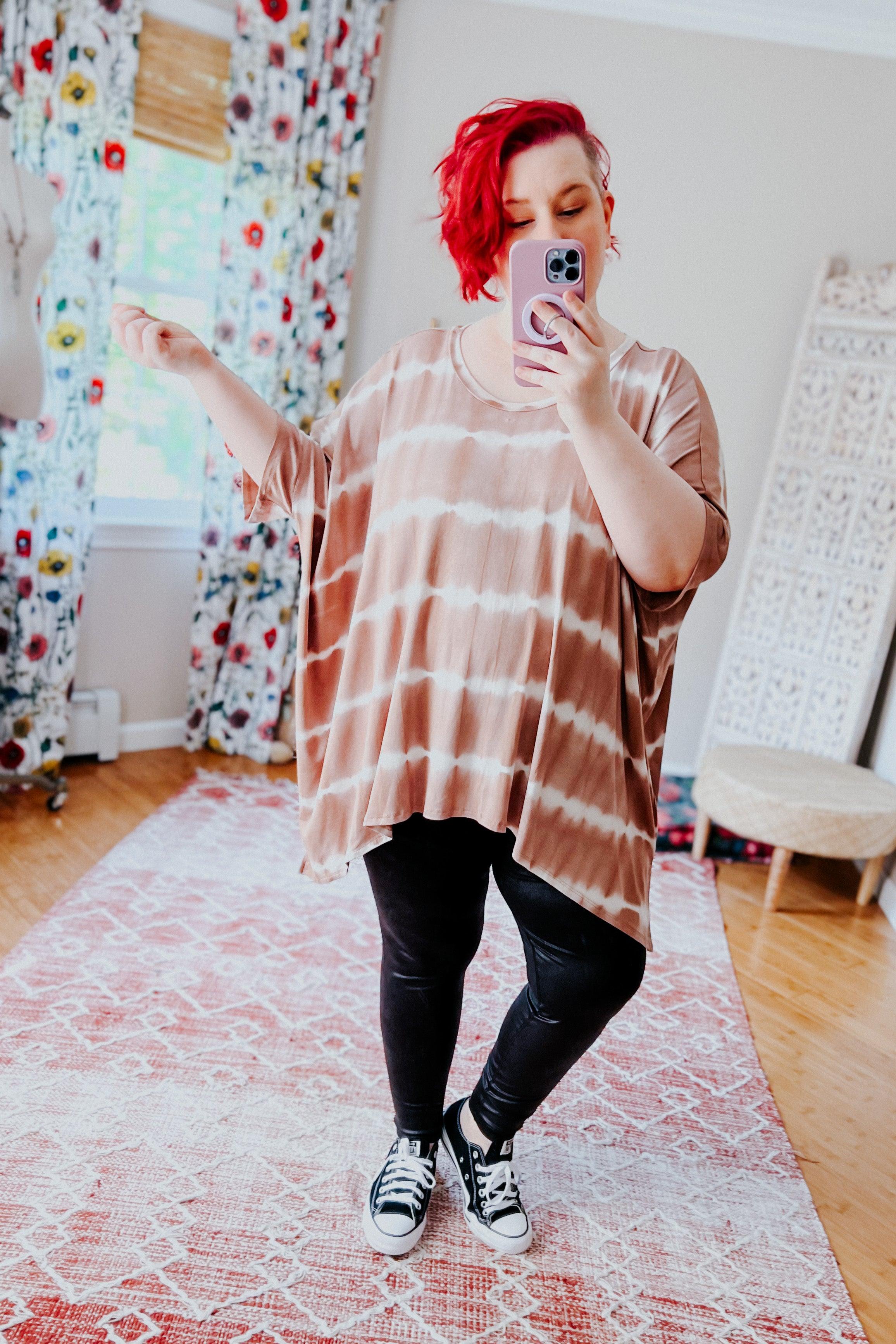 Woman with bright red hair wearing a loose striped tie dye tunic and black leggings taking a mirror selfie indoors