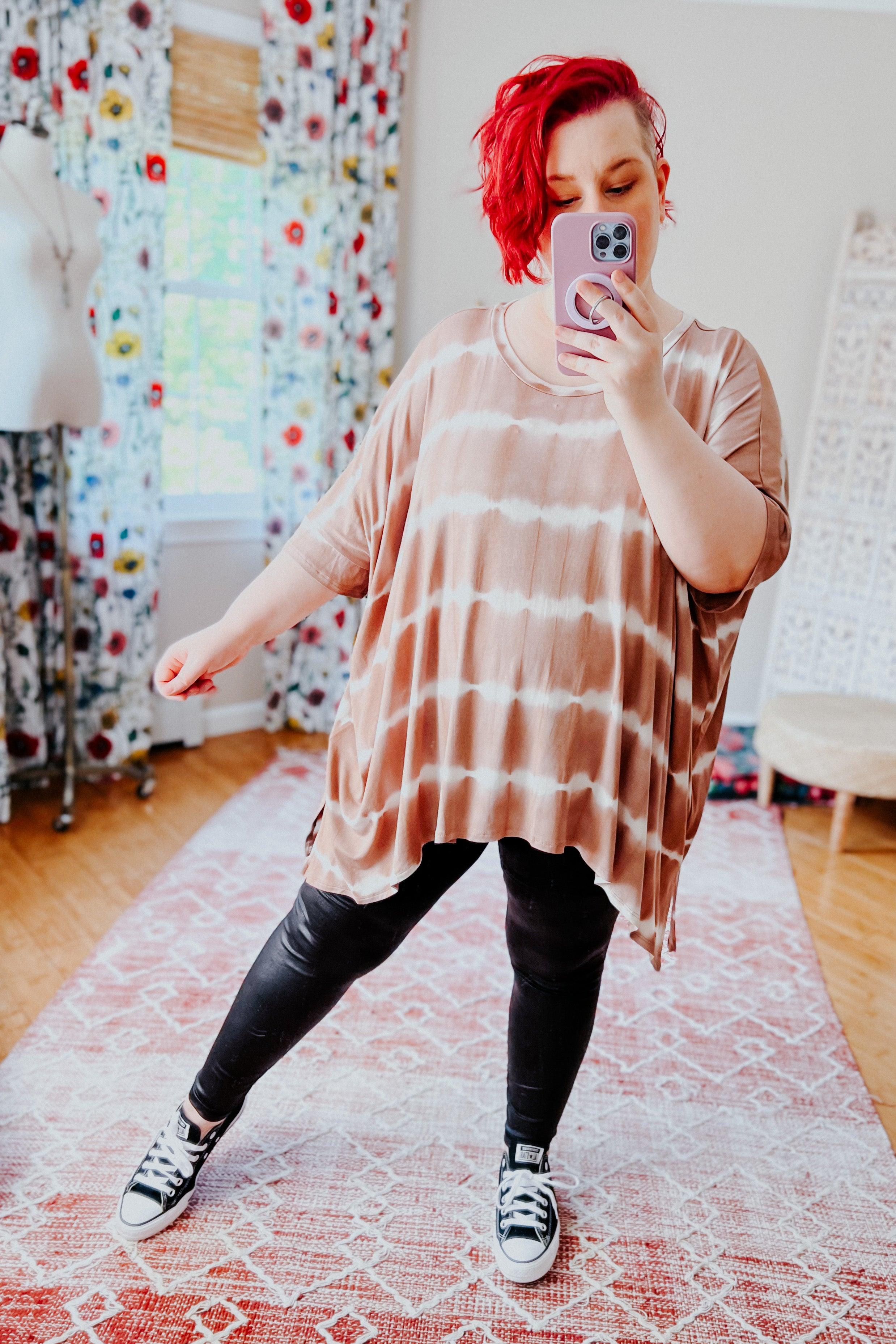 Woman with bright red hair wearing a loose striped tie dye tunic and black pants taking a selfie indoors