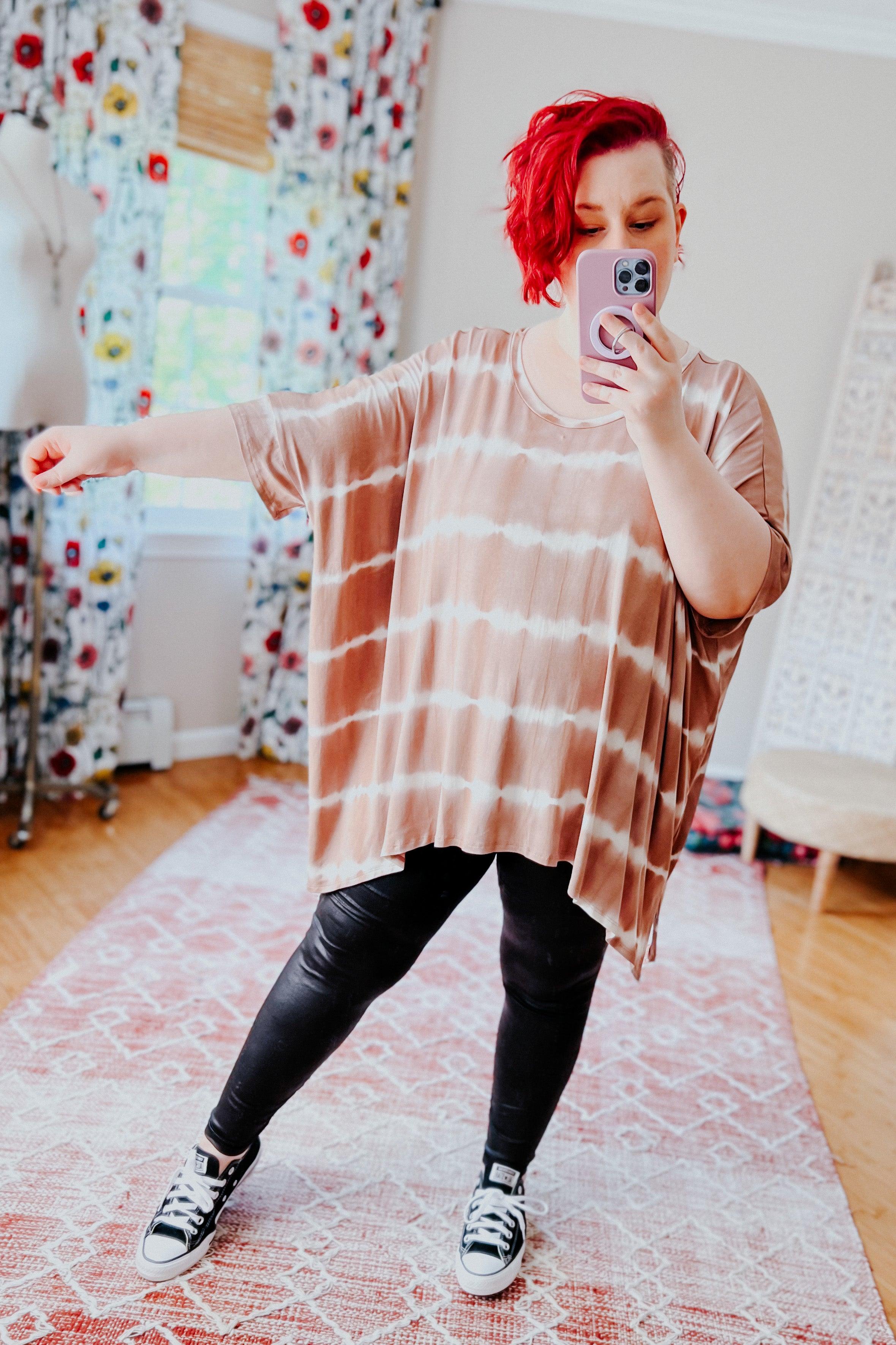 Woman with bright red hair wearing a striped tie dye tunic and black leggings taking a mirror selfie in a cozy room