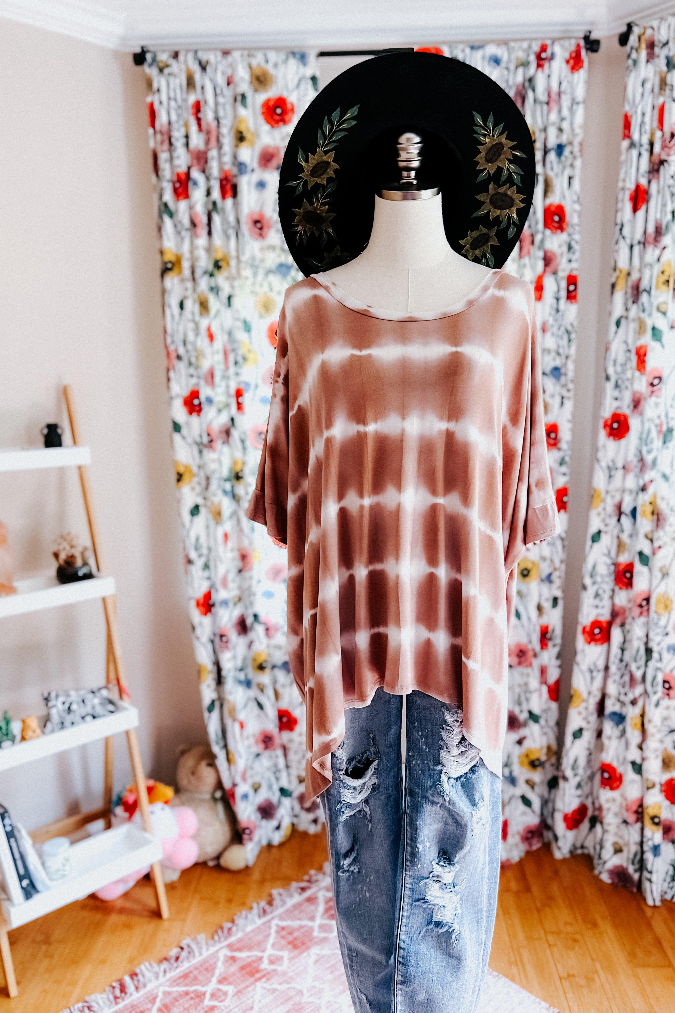 Mannequin dressed in a pink striped tie dye tunic with distressed blue jeans and a black floral wide-brim hat indoors