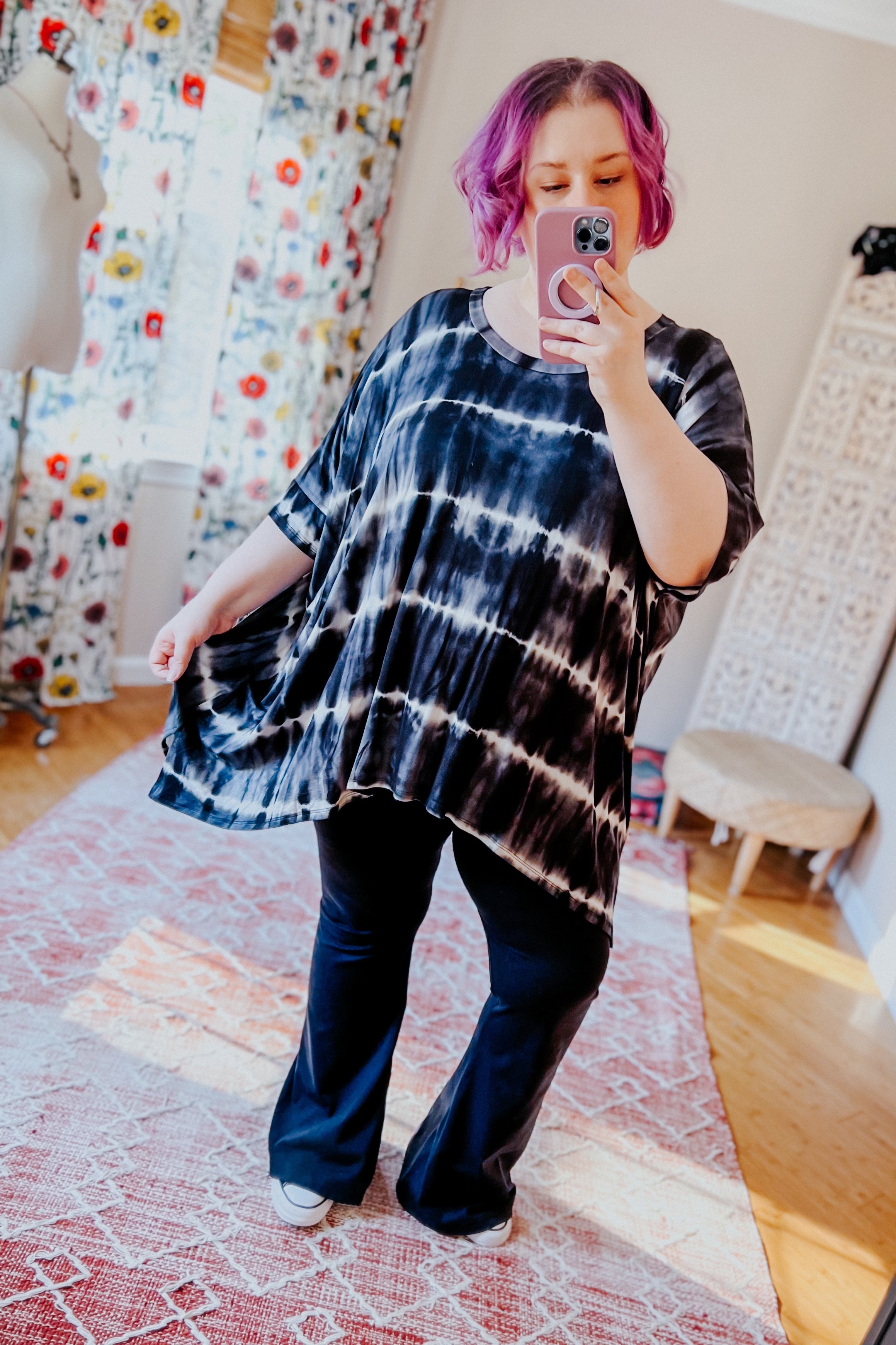 Woman with purple hair wearing a loose black striped tie dye tunic and black pants taking a mirror selfie in a bright room
