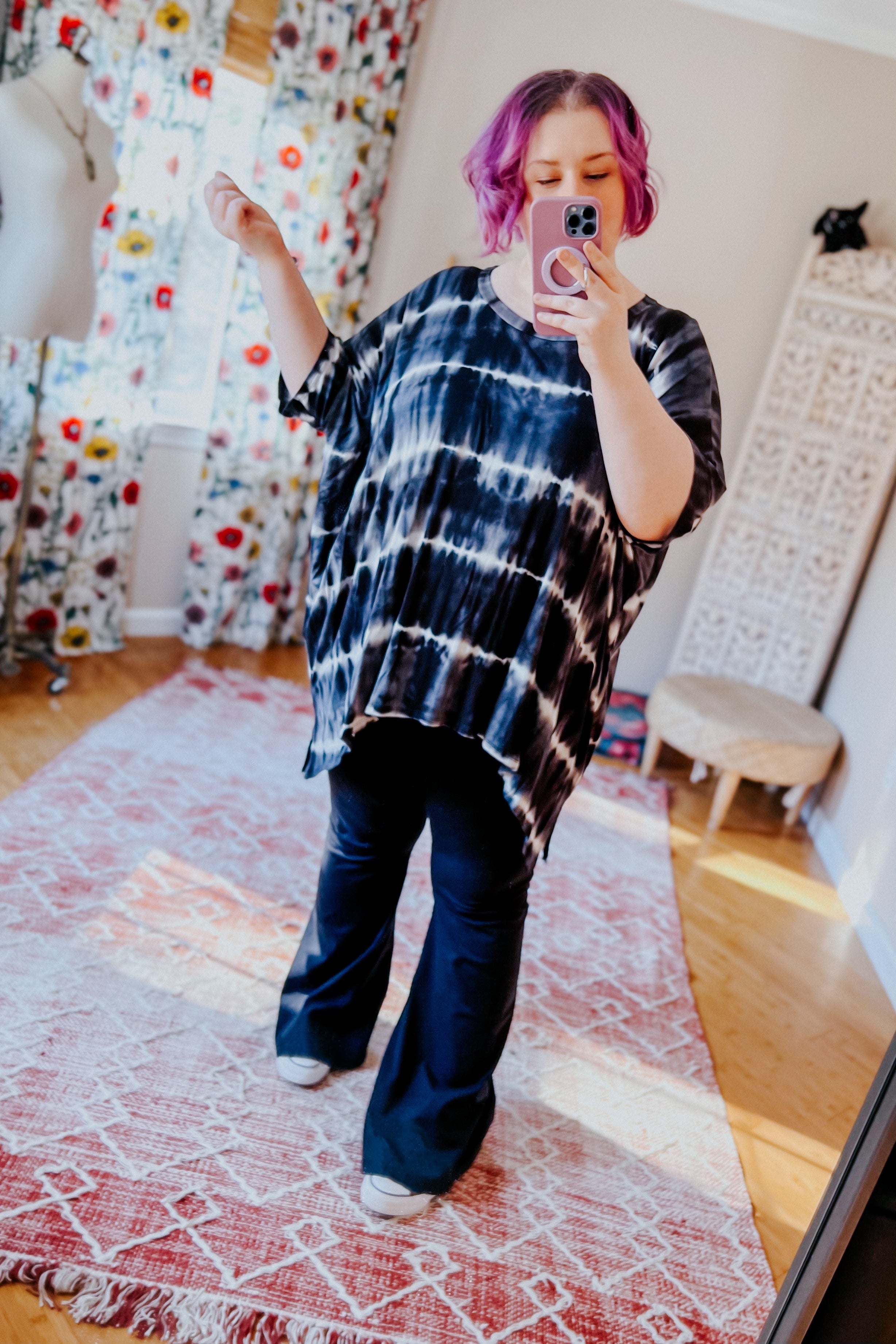 Woman with purple hair wearing a loose black striped tie dye tunic and black pants taking a mirror selfie indoors