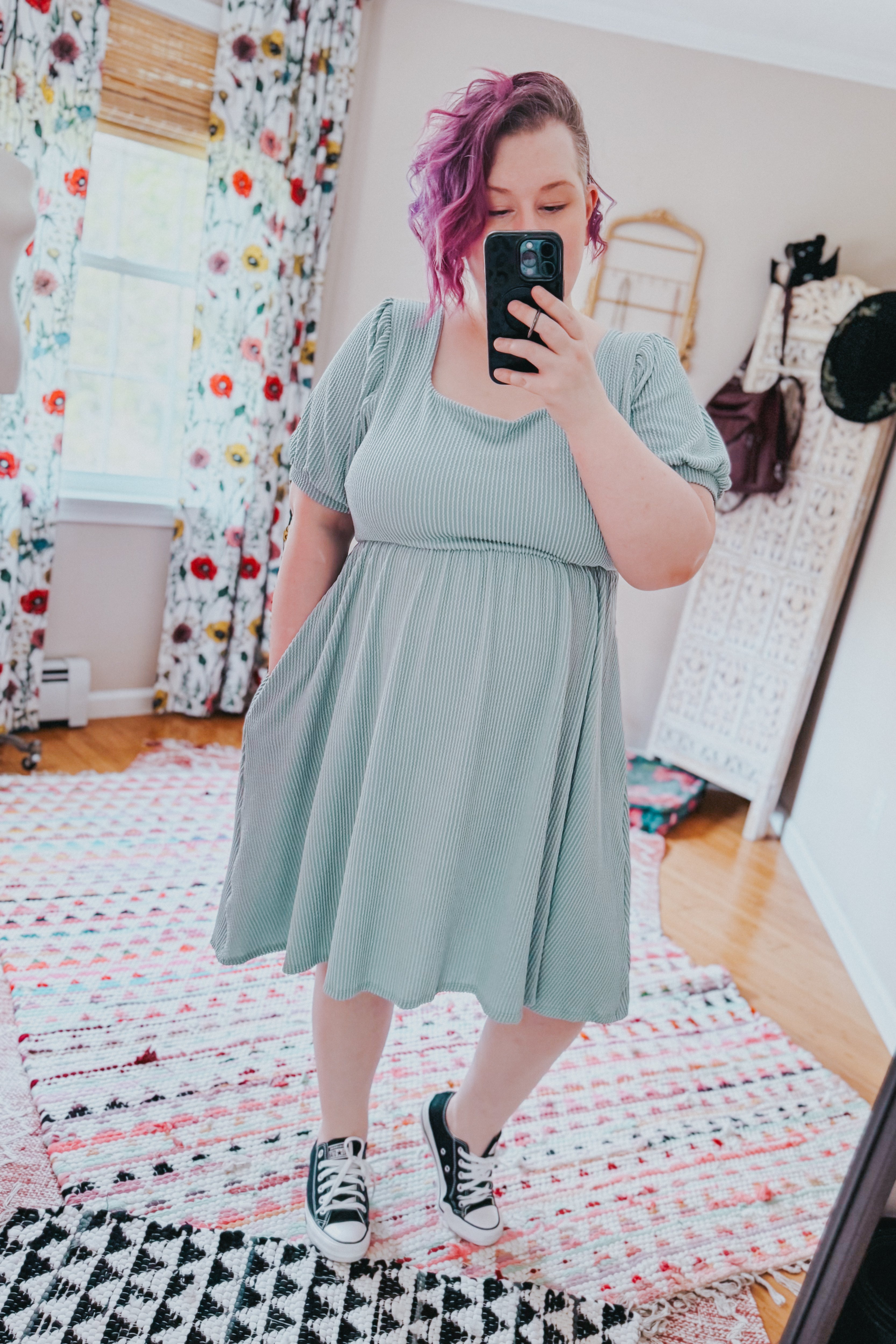 Woman with purple hair wearing a light sage ribbed knit dress and black sneakers taking a mirror selfie indoors