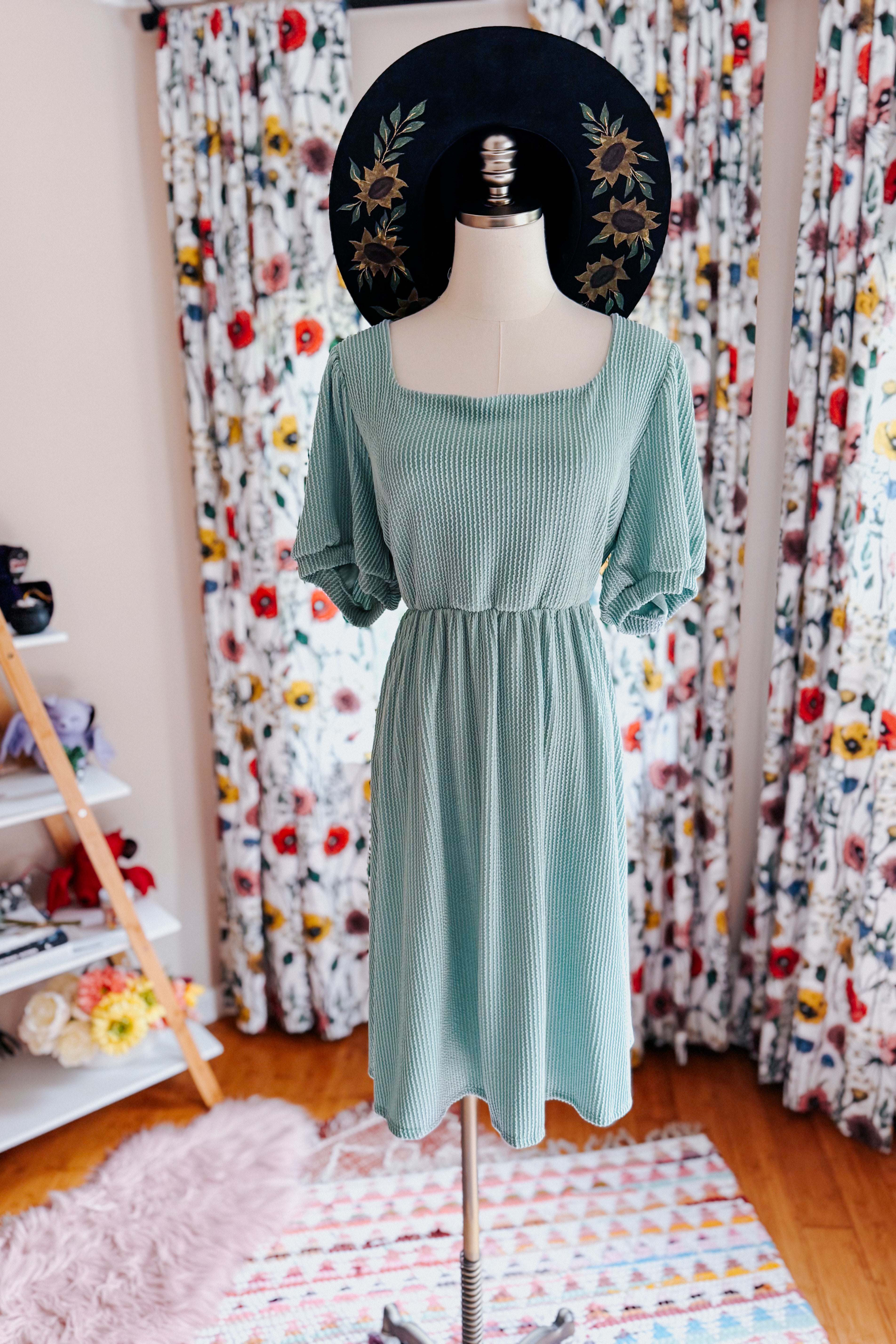 Light sage cable ribbed knit dress with puff sleeves displayed on a mannequin in a cozy, floral-decor room