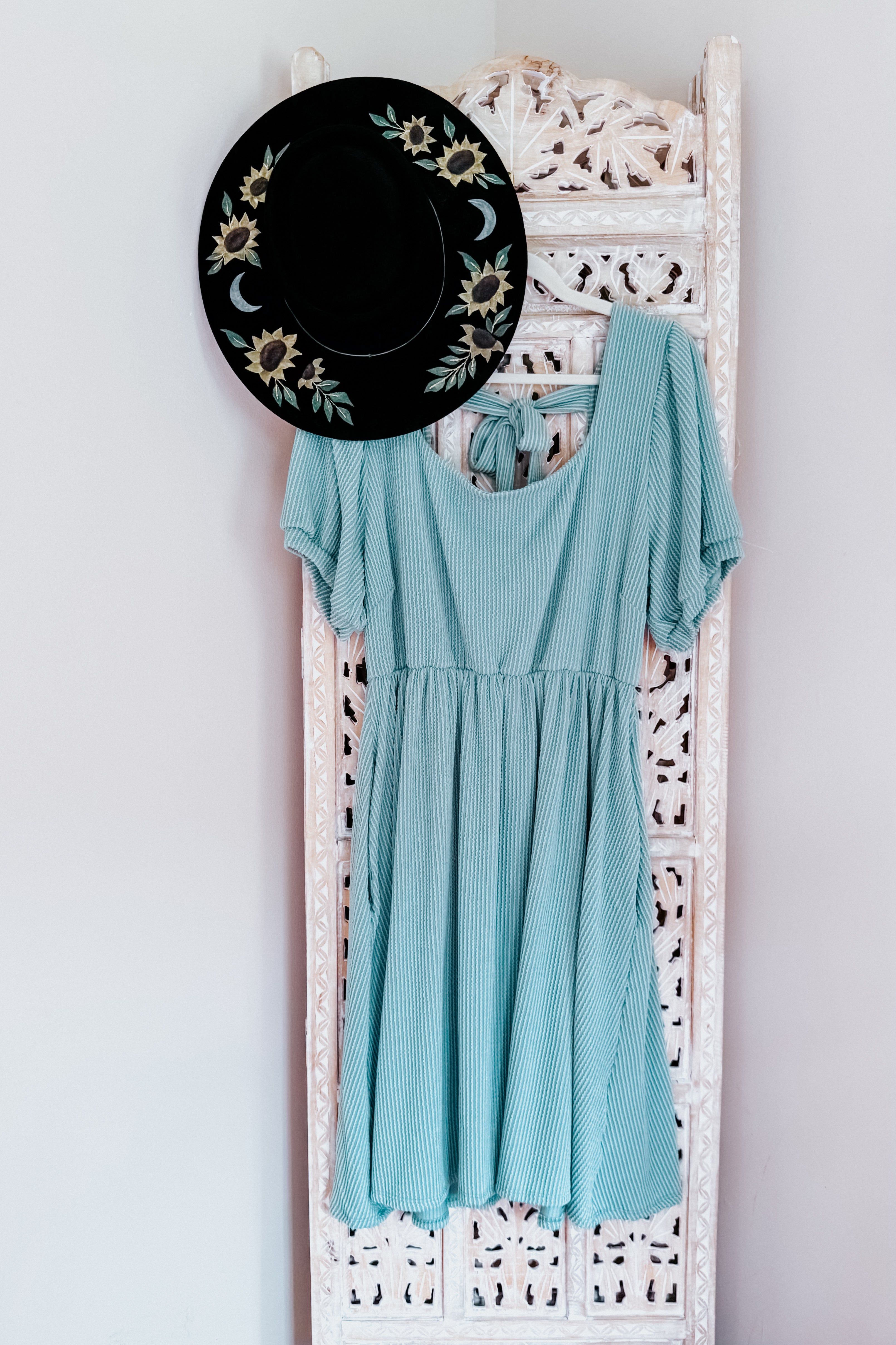 Light sage cable ribbed knit dress hanging on carved white wooden panel with black hat featuring sunflowers and moons