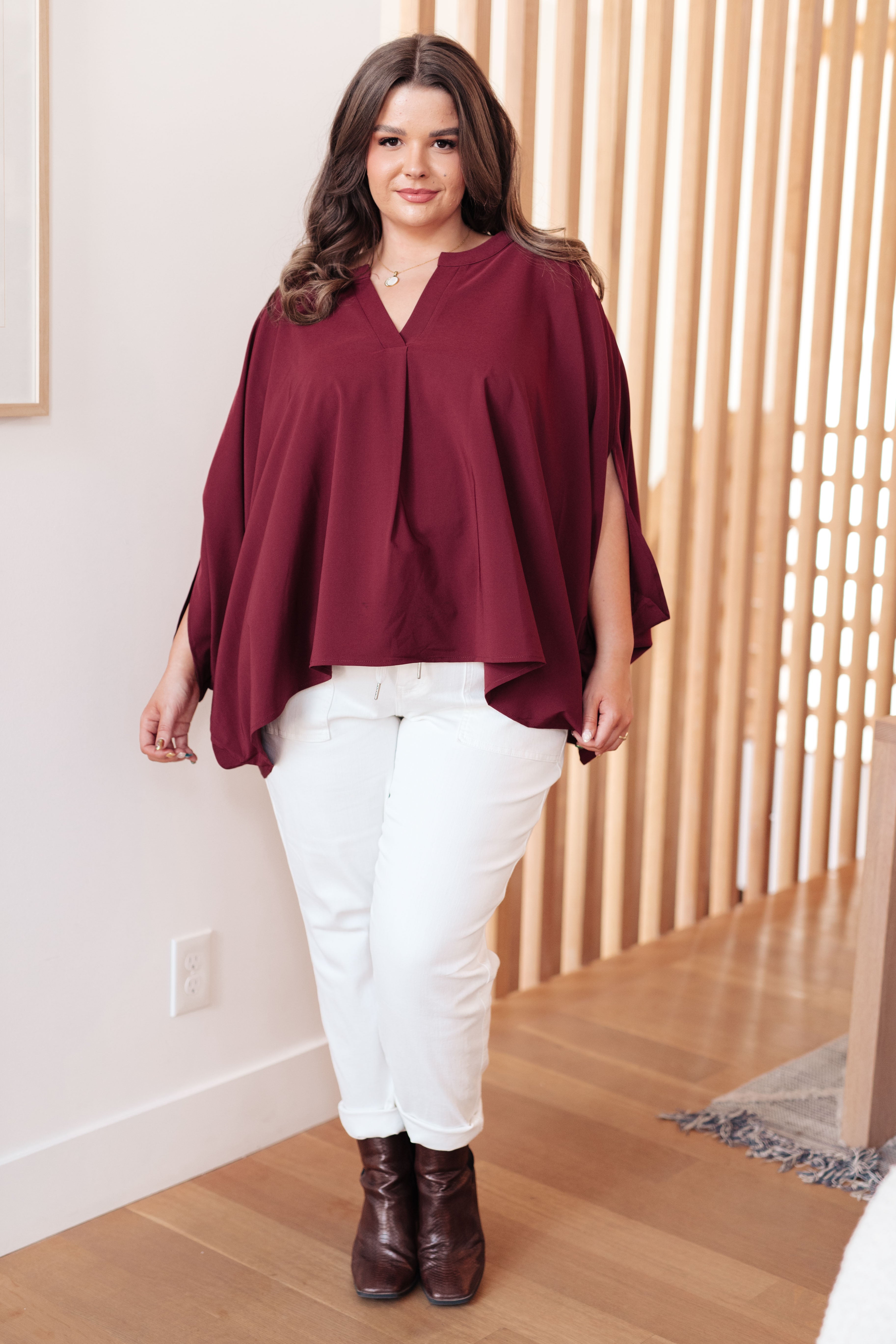 Plus size model wearing a wine-colored flowy blouse with slit sleeves paired with white jeans and dark boots indoors
