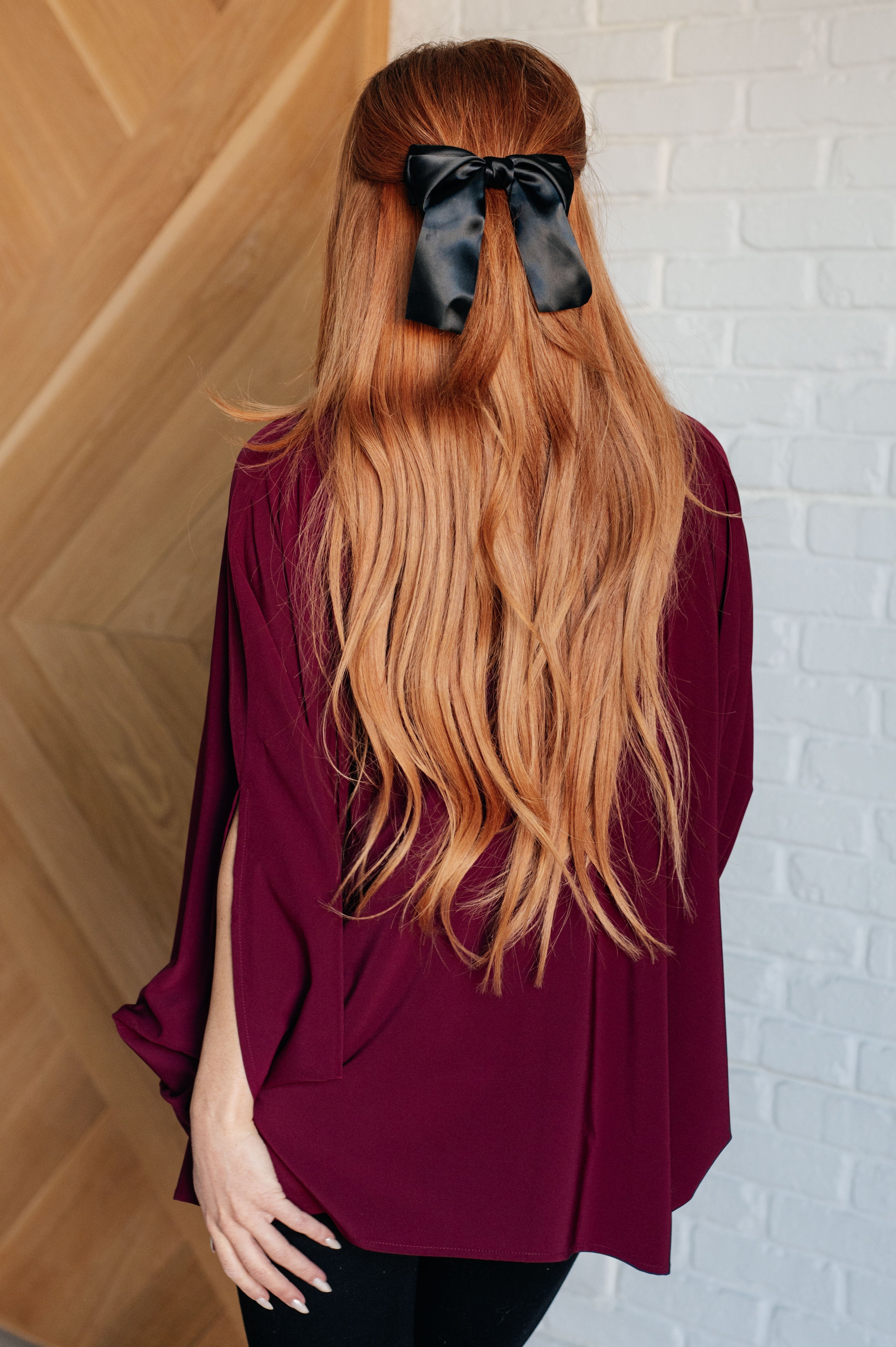 Woman with long red hair wearing a wine-colored blouse and black satin bow standing against wood and brick walls