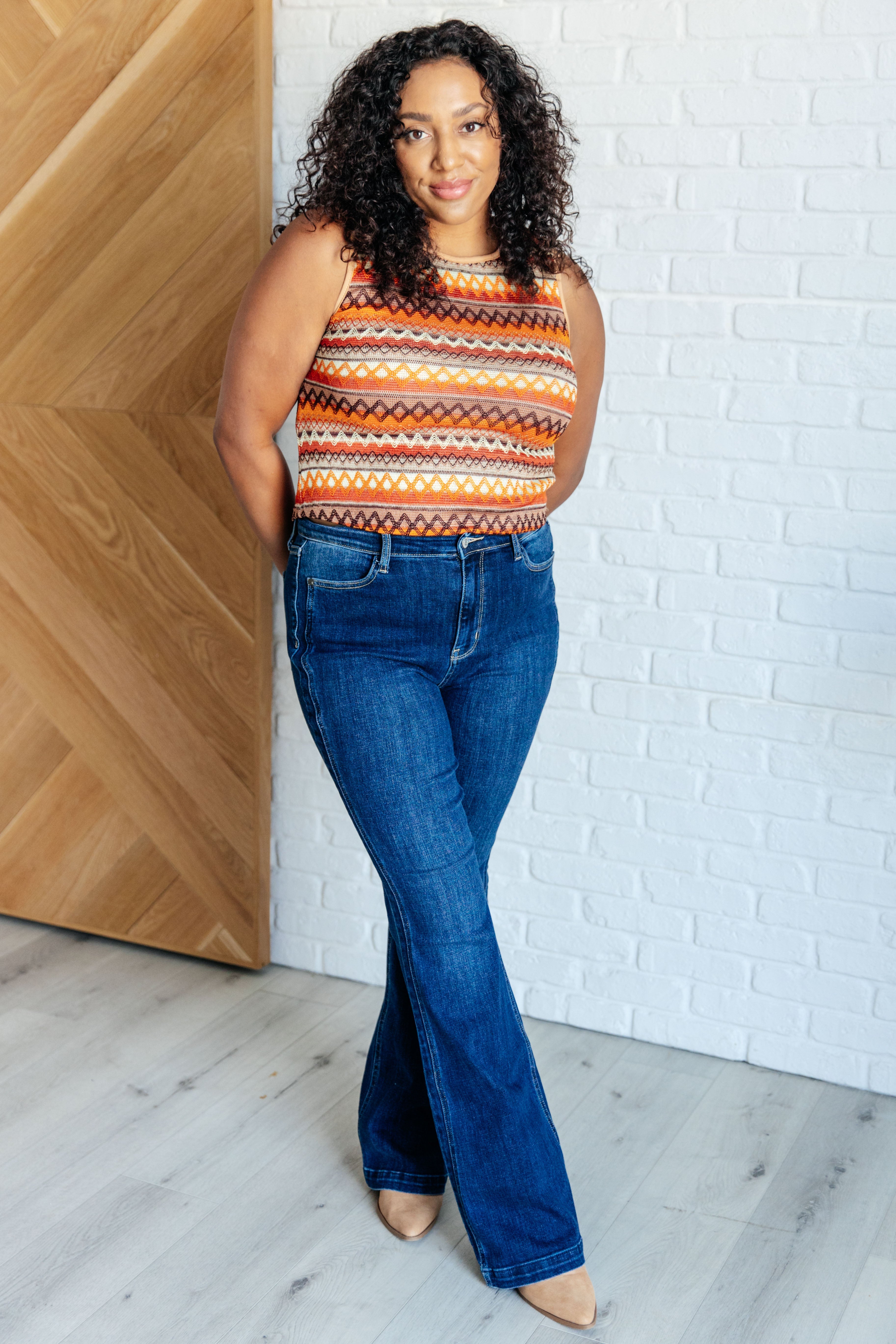 Woman wearing a patterned orange knit tank paired with dark flared jeans and nude heels indoors by a white brick wall