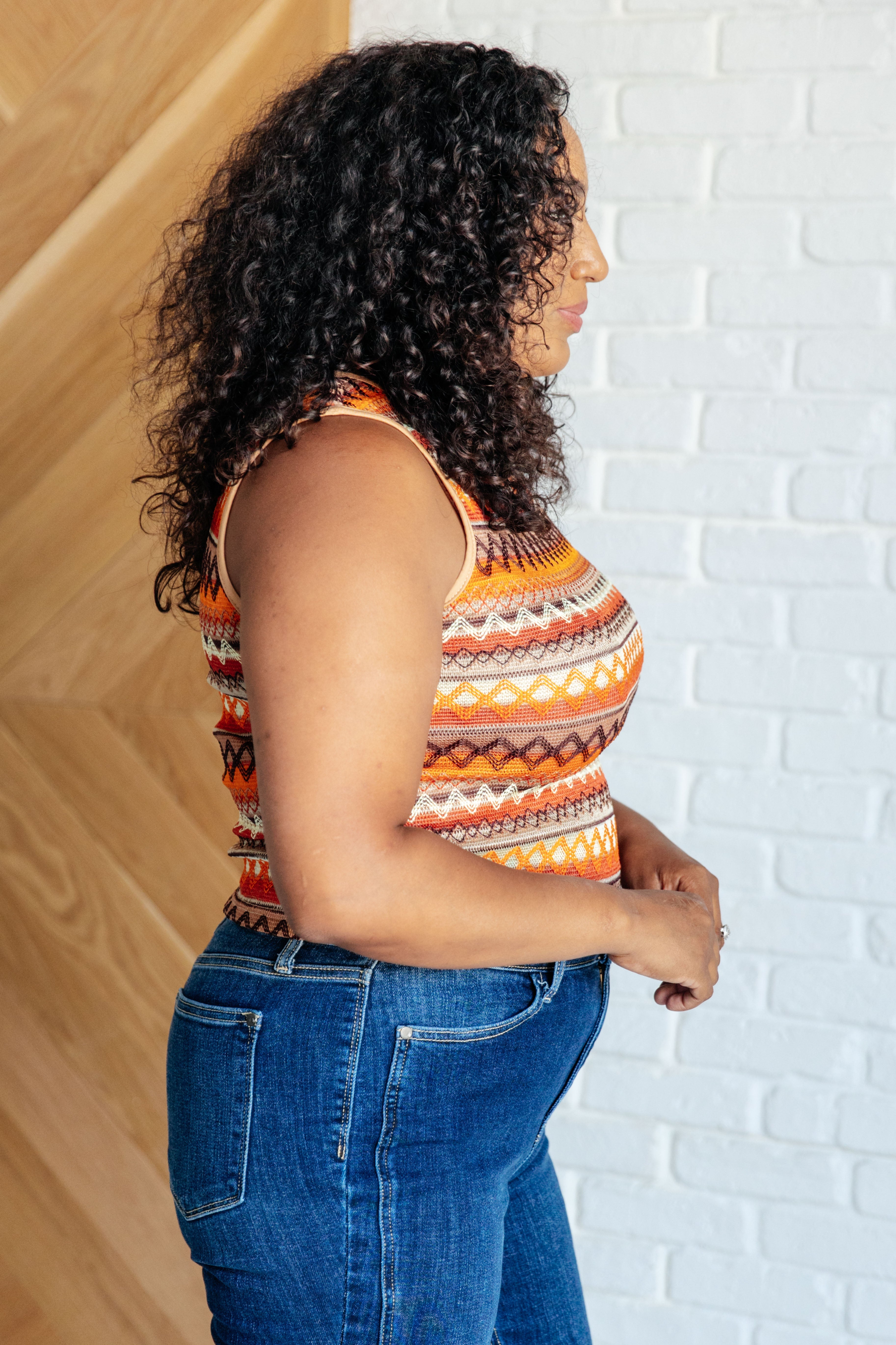Curly haired woman wearing a vibrant orange patterned knit tank paired with blue high-rise jeans against a white wall