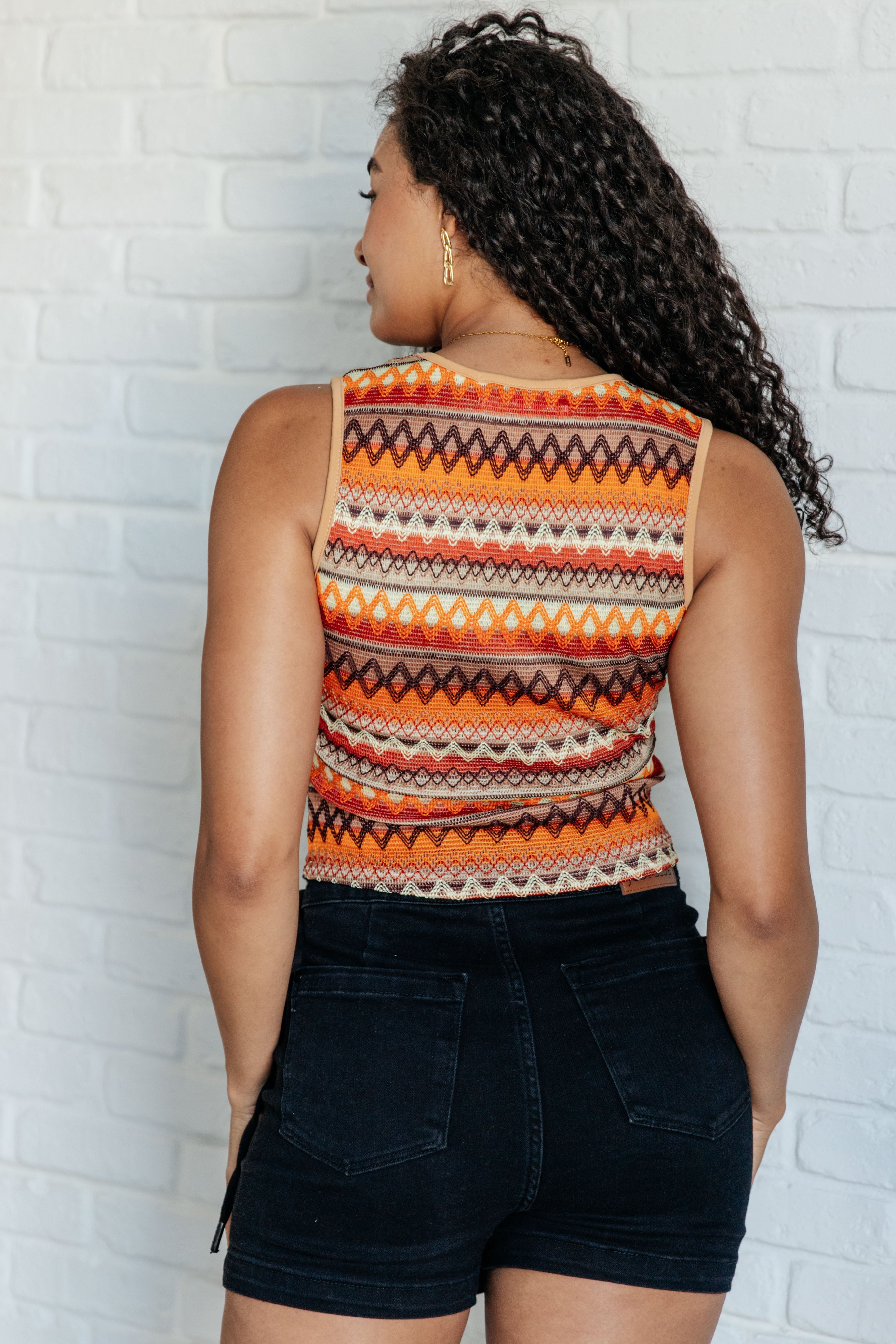 Woman wearing an orange patterned knit tank top paired with black high-waisted shorts against white brick wall