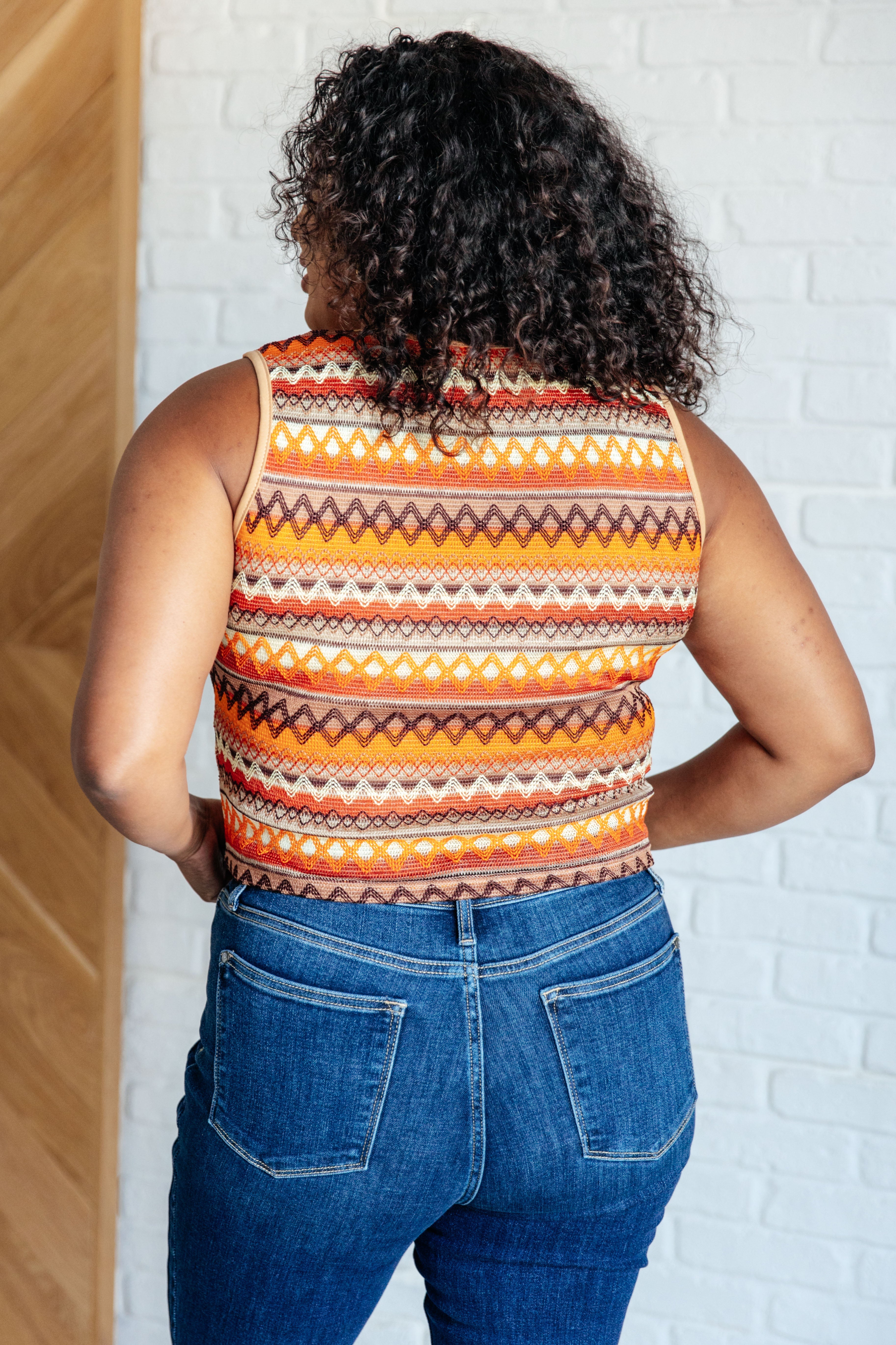 Curly-haired woman wearing an eclectic orange patterned knit tank and blue jeans standing indoors with hands on hips
