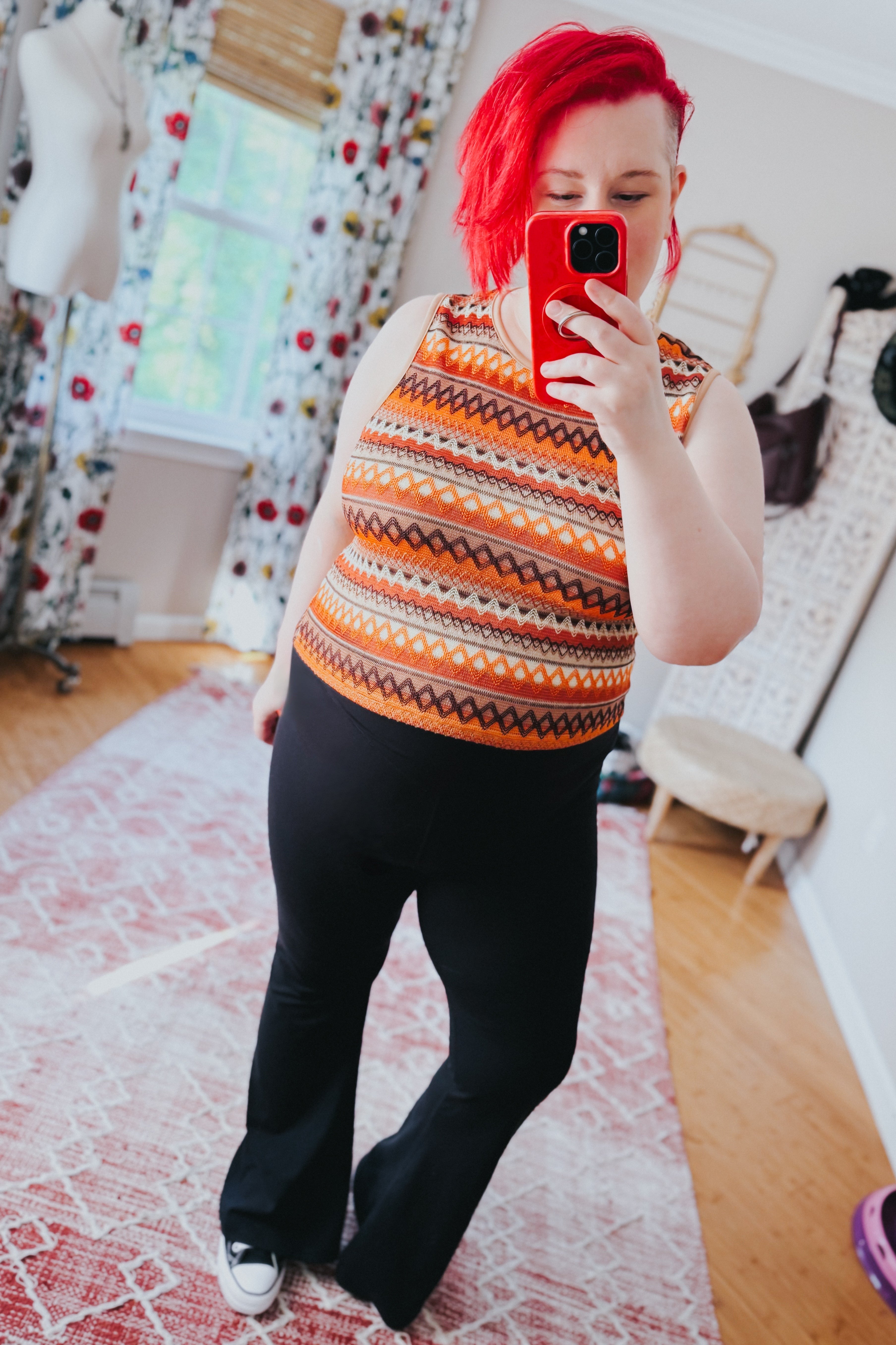 Woman with bright red hair wearing an orange patterned knit tank top paired with black pants and sneakers in a cozy room