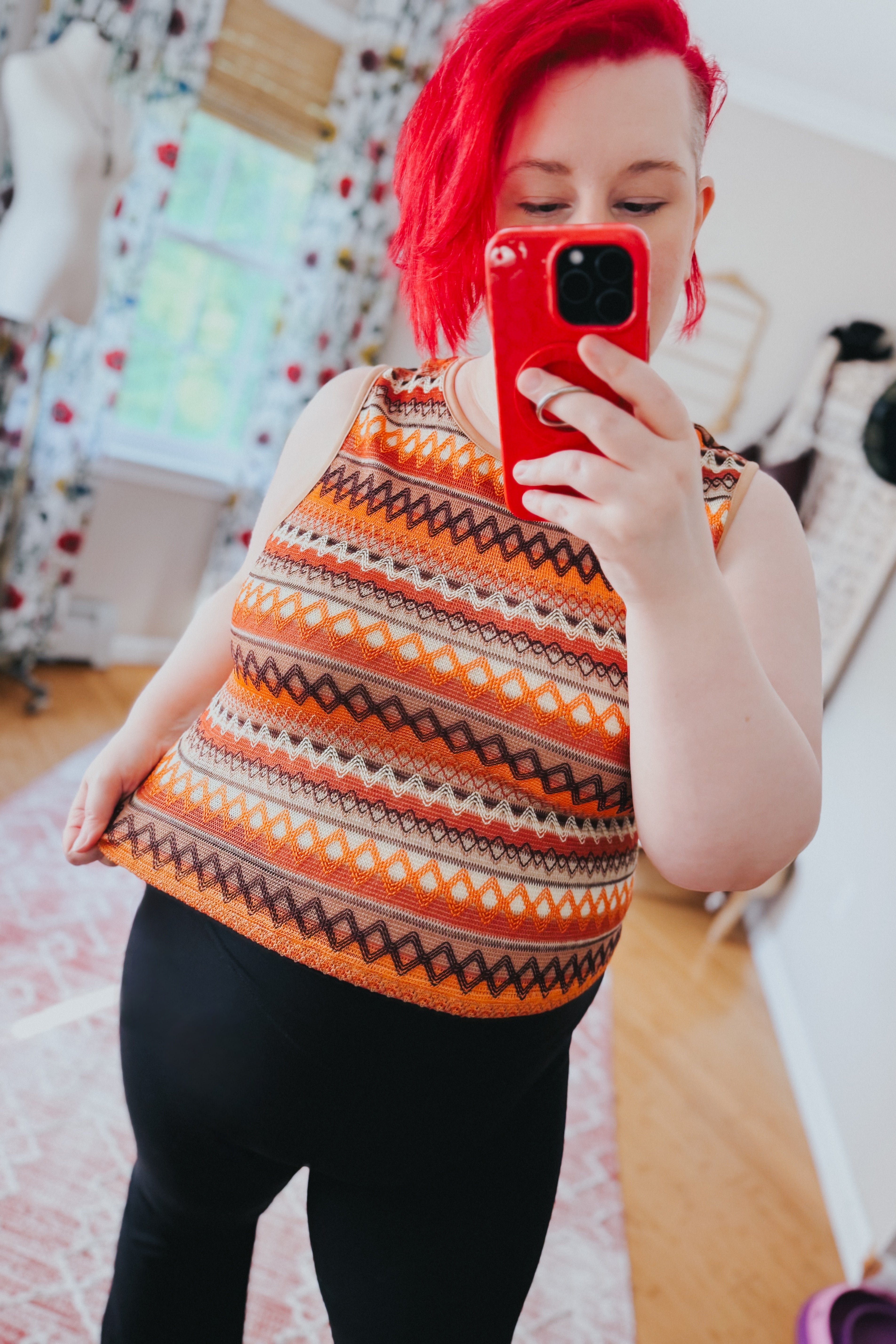 Woman with bright red hair wearing an orange patterned knit tank top and black pants taking a mirror selfie indoors