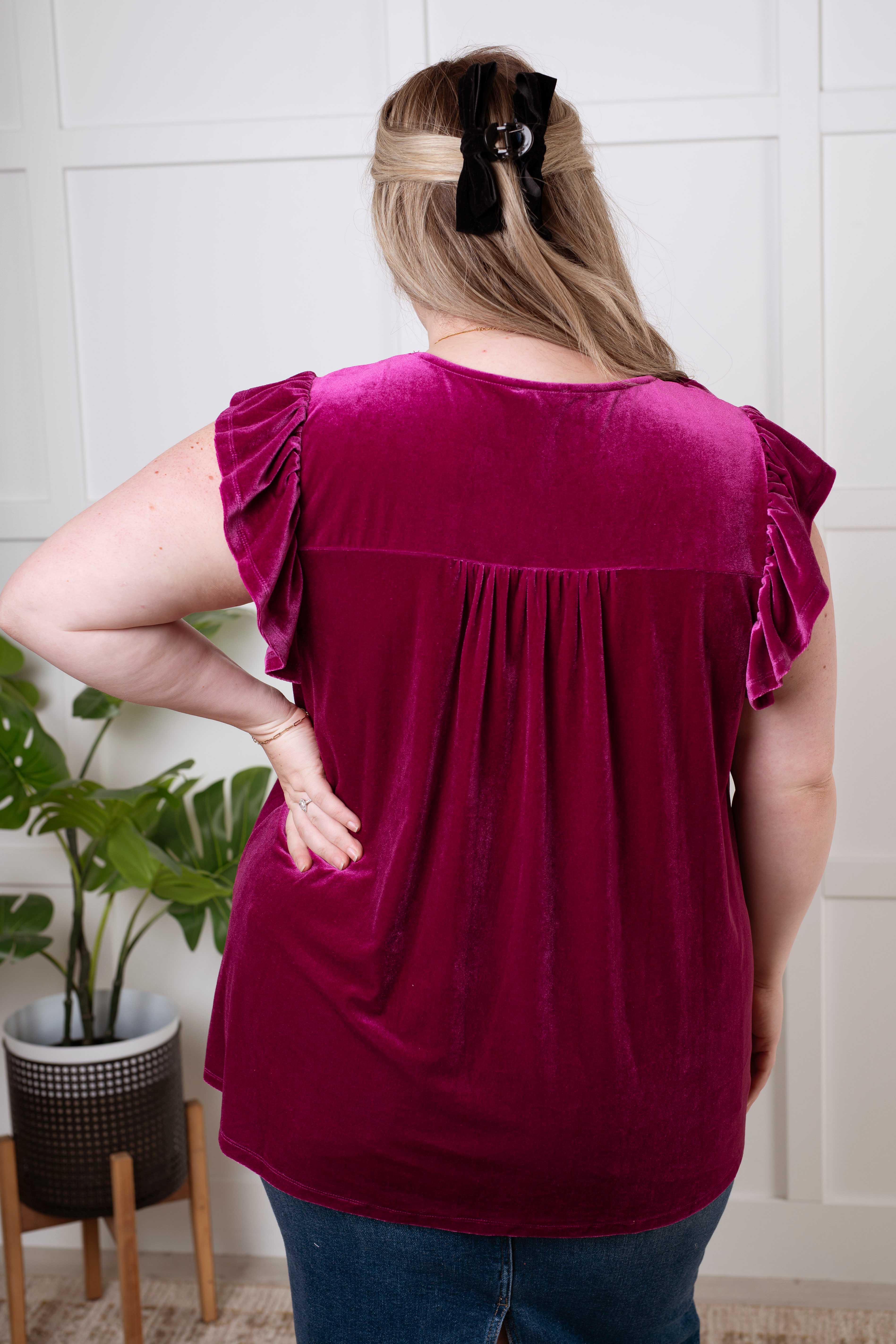 Back view of woman wearing magenta velveteen blouse with ruffled sleeves, standing indoors near a plant and white paneled wall